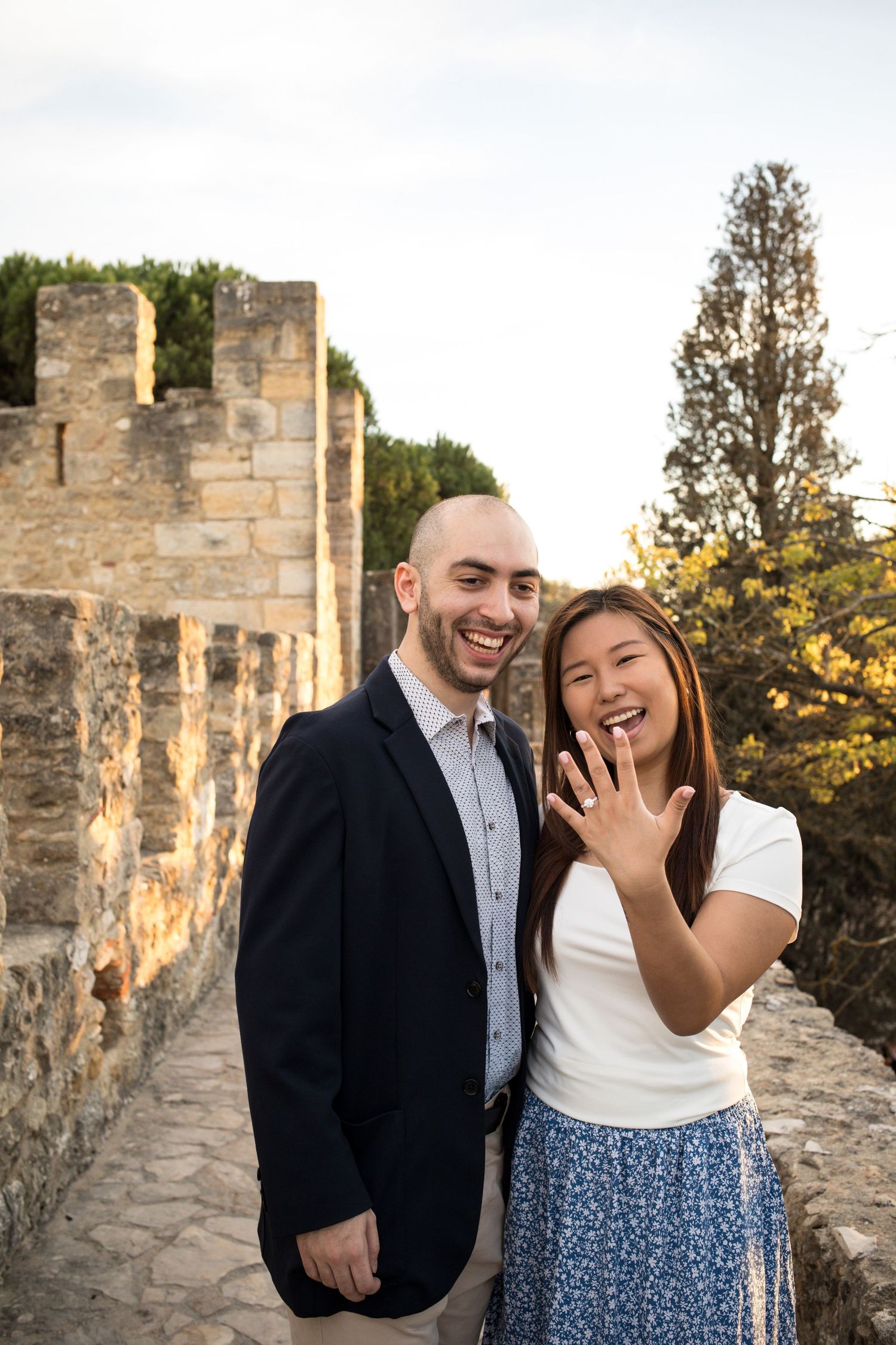 Engaged couple laughs joyfully as woman shows off engagement ring at historic stone castle.