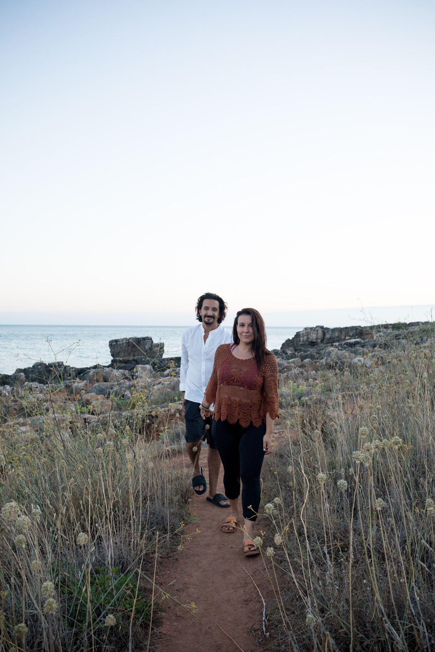 Couple walking hand in hand along a coastal path surrounded by wild grass with ocean in background.