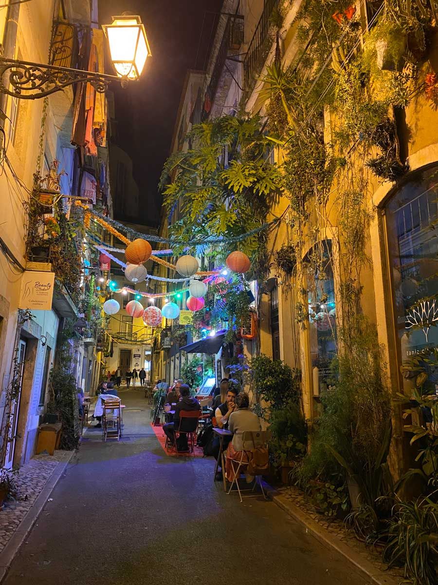 Charming narrow alley at night with colorful paper lanterns, lush plants, and outdoor dining in a European city.