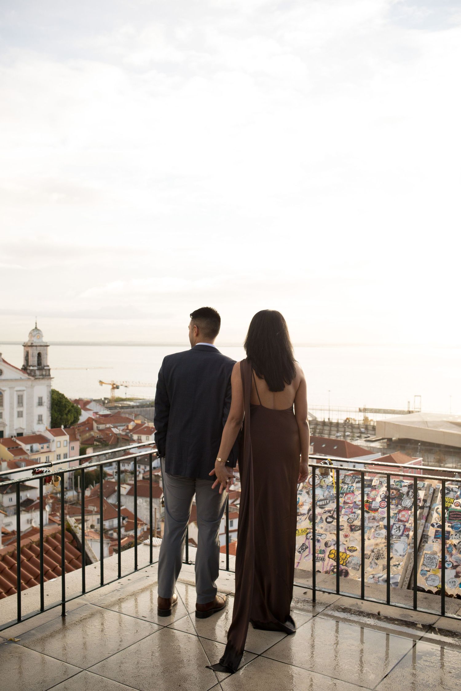 Couple holding hands on a balcony overlooking a coastal city at sunset.