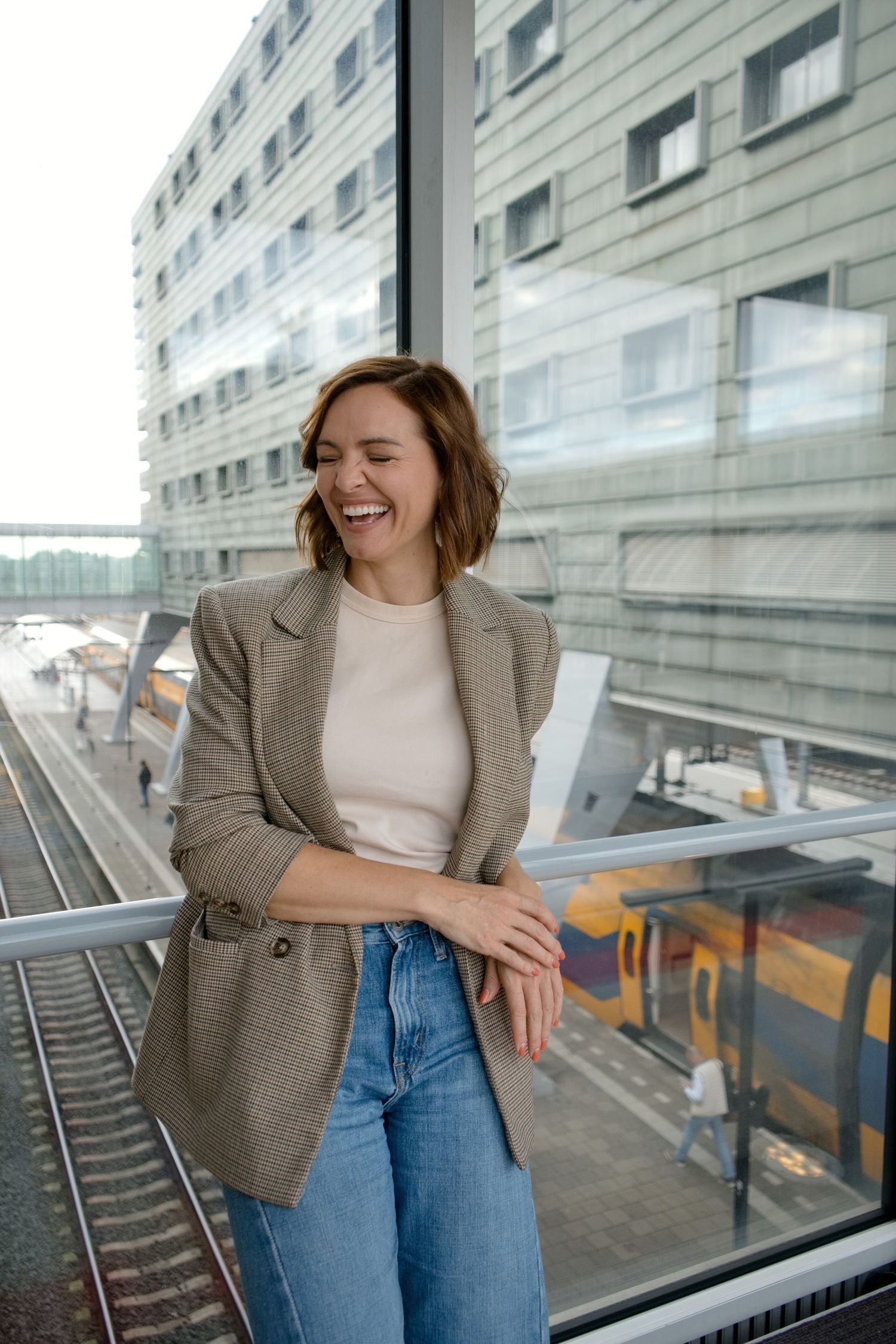Smiling woman in a plaid blazer and jeans laughs while leaning on a glass railing above train tracks at a modern railway station.