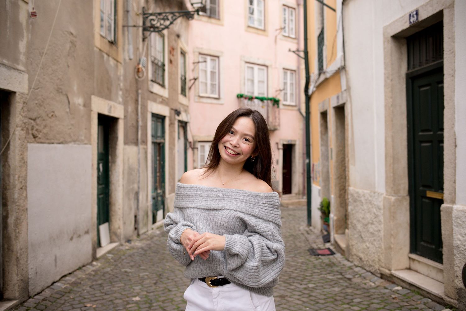Smiling young woman in a gray off-shoulder sweater posing on a charming cobblestone street.