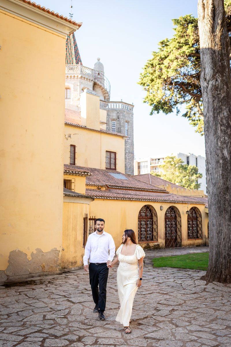 Couple holding hands walking along cobblestone path beside yellow historic building on sunny day.