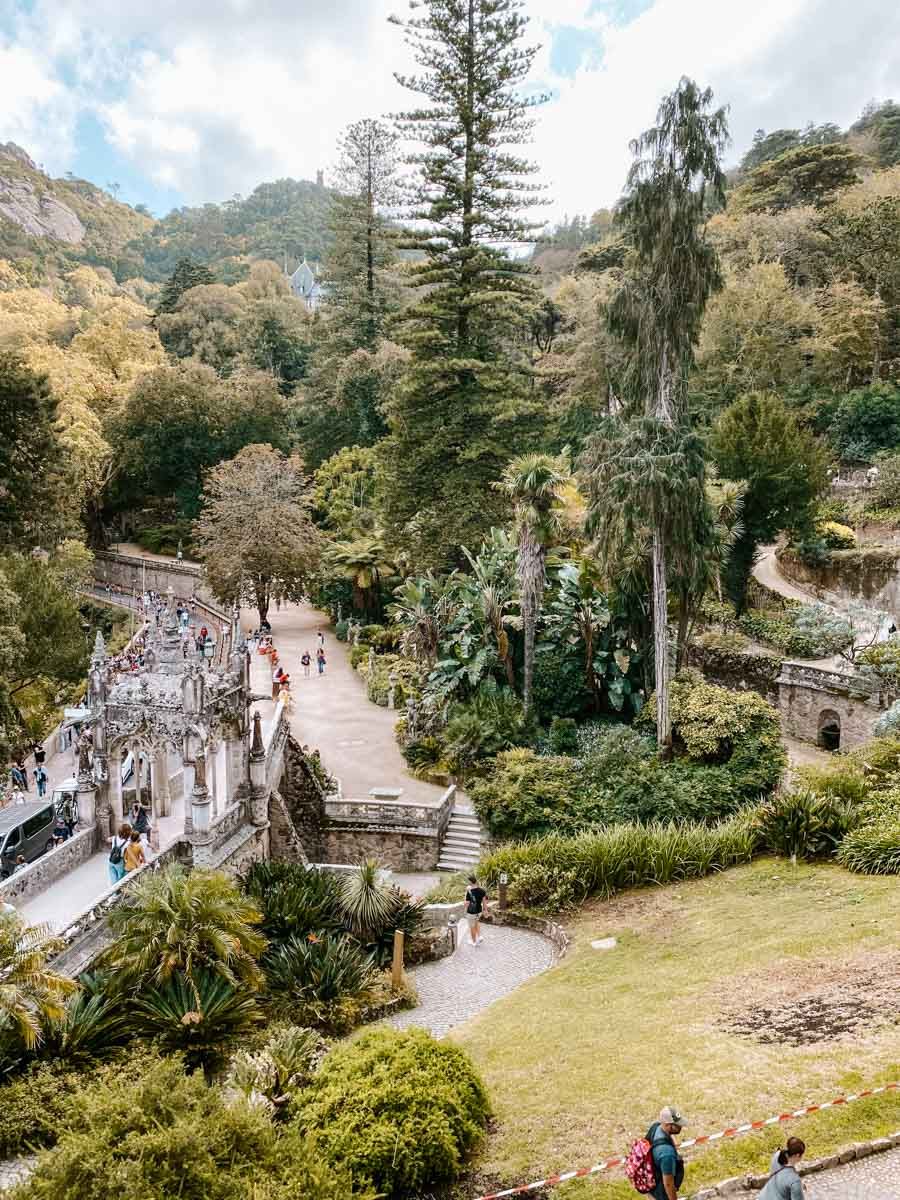 Lush garden landscape with stone pathways, tall pine trees, and visitors exploring terraced greenery on a cloudy day.
