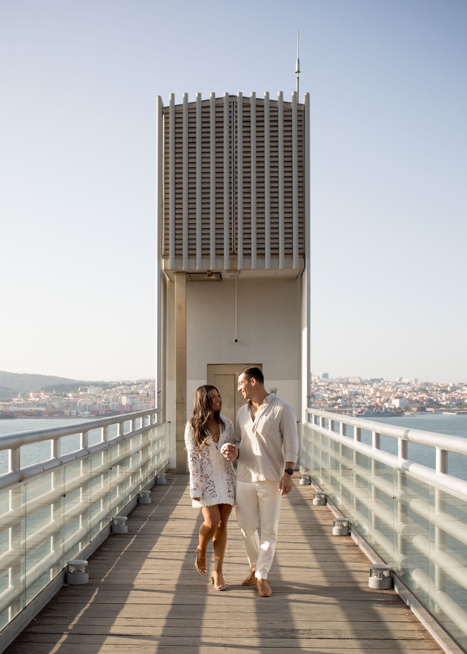 Couple walking hand in hand on a bridge walkway with a city skyline and tower in the background.