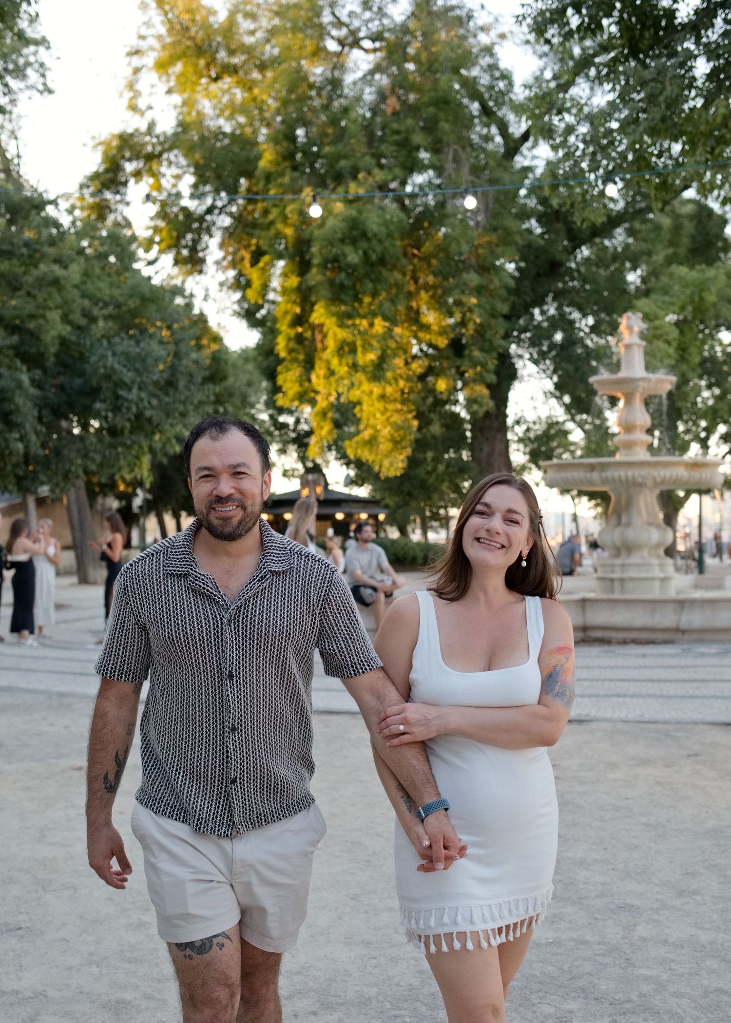 Couple holding hands and smiling while walking in a sunny park with a fountain and trees in the background.