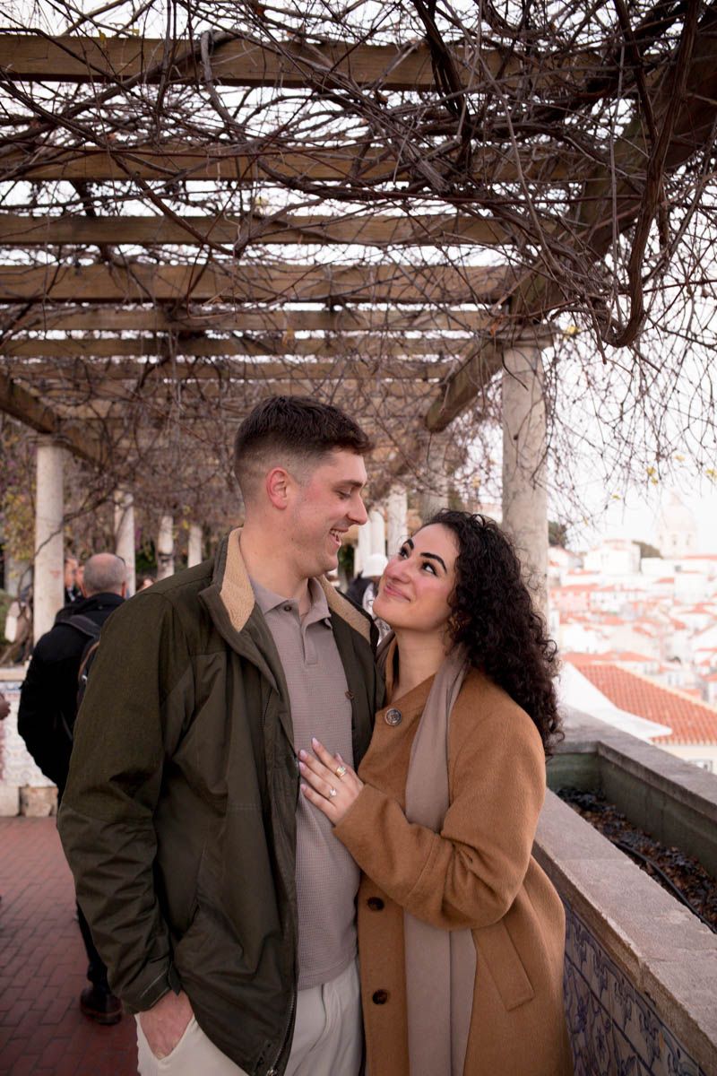 Couple sharing a romantic moment under a pergola with bare vines, woman in camel coat smiling at man in green jacket.