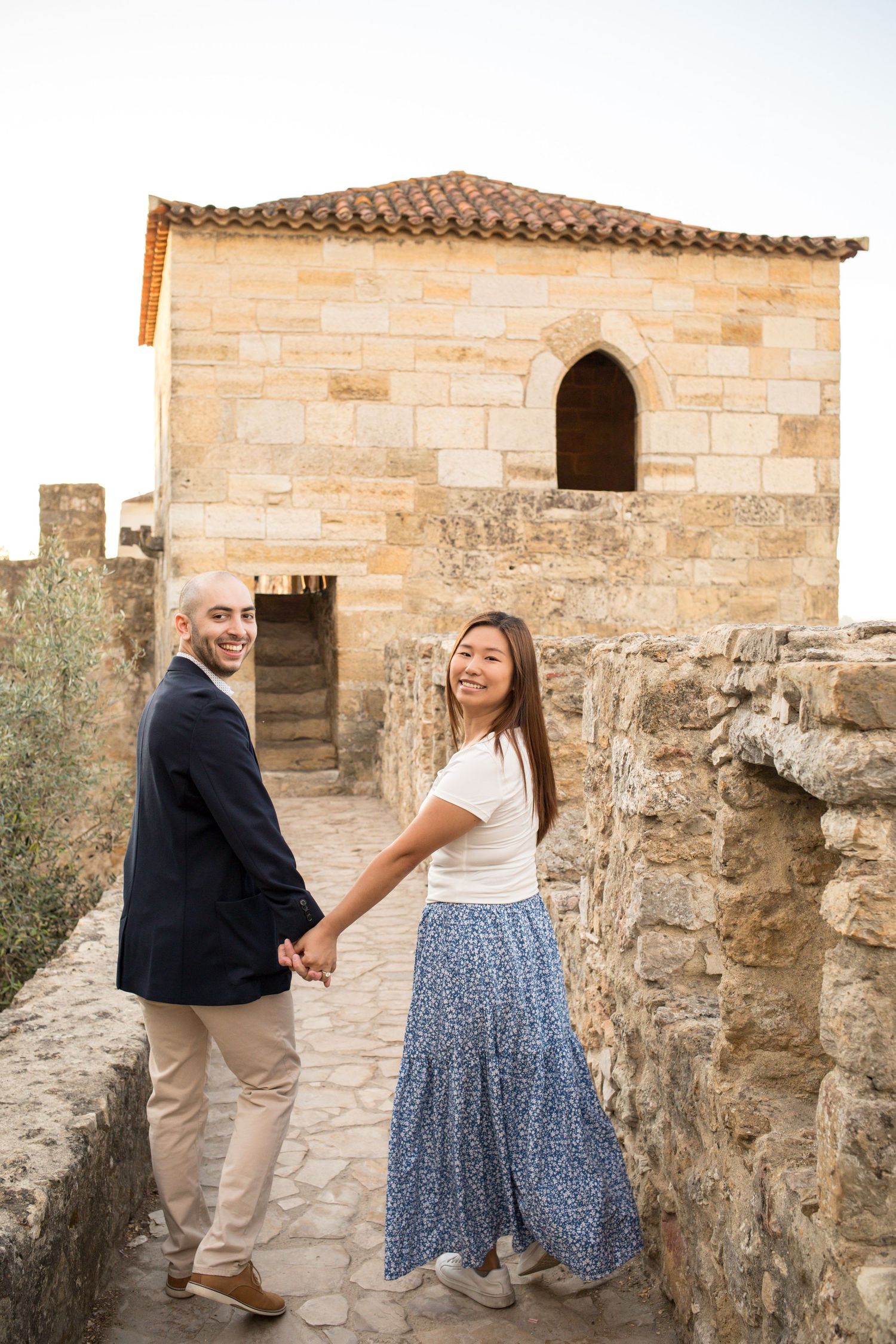 Couple holding hands smiling on ancient stone castle walkway with historic building backdrop.