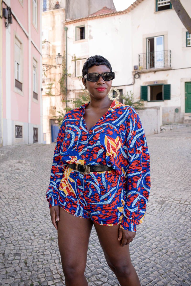 Woman in vibrant African print co-ord set with sunglasses posing on cobblestone street in historic European town.