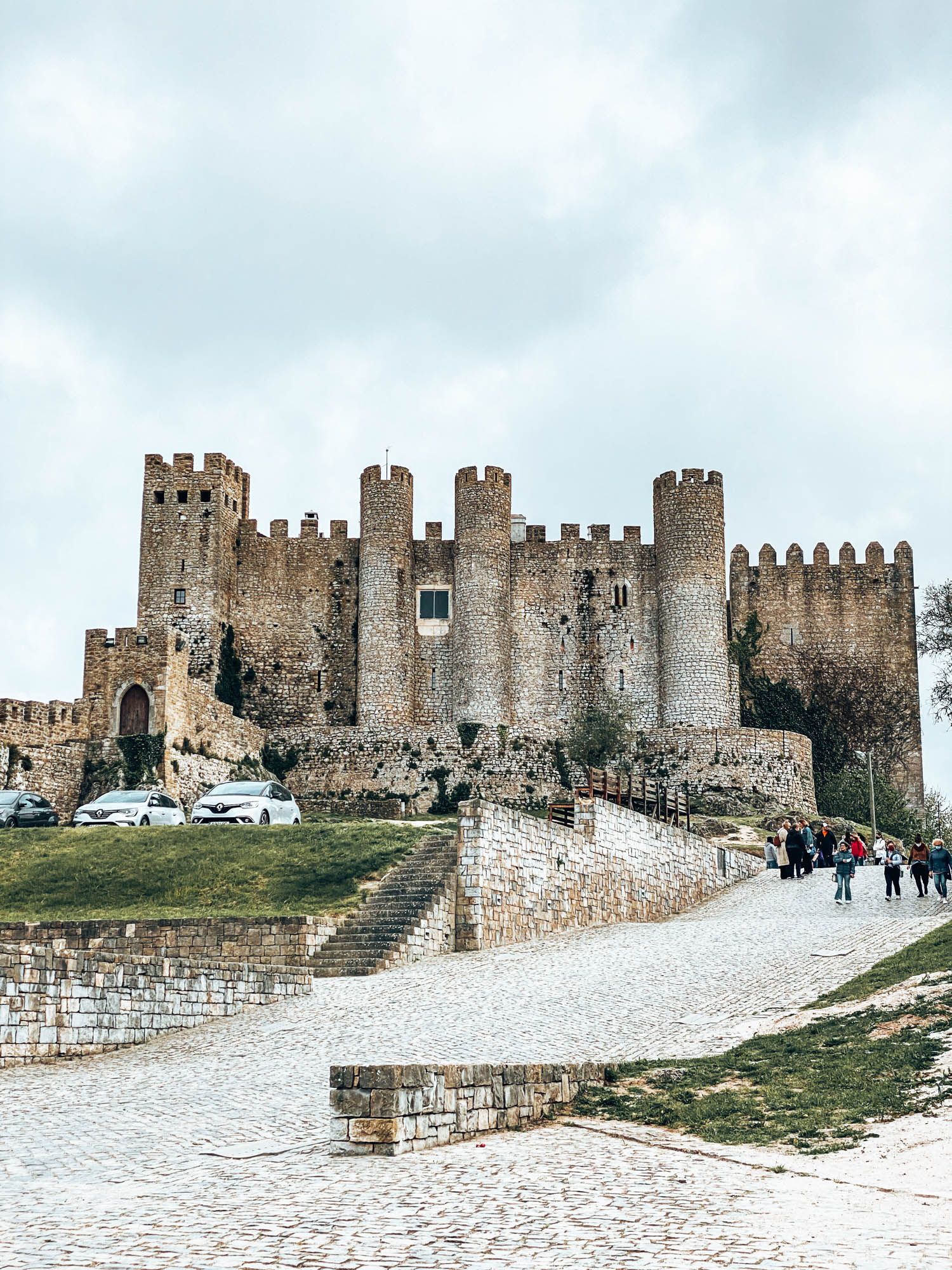 Medieval Óbidos Castle in Portugal with stone towers, battlements, and cobblestone path under cloudy skies.