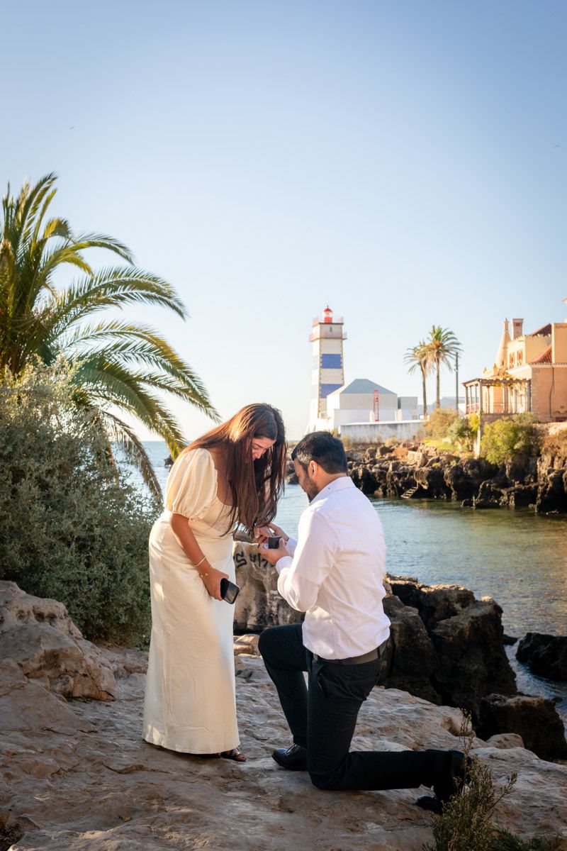 Man proposing to woman on rocky seaside with lighthouse and palm trees in background at golden hour.