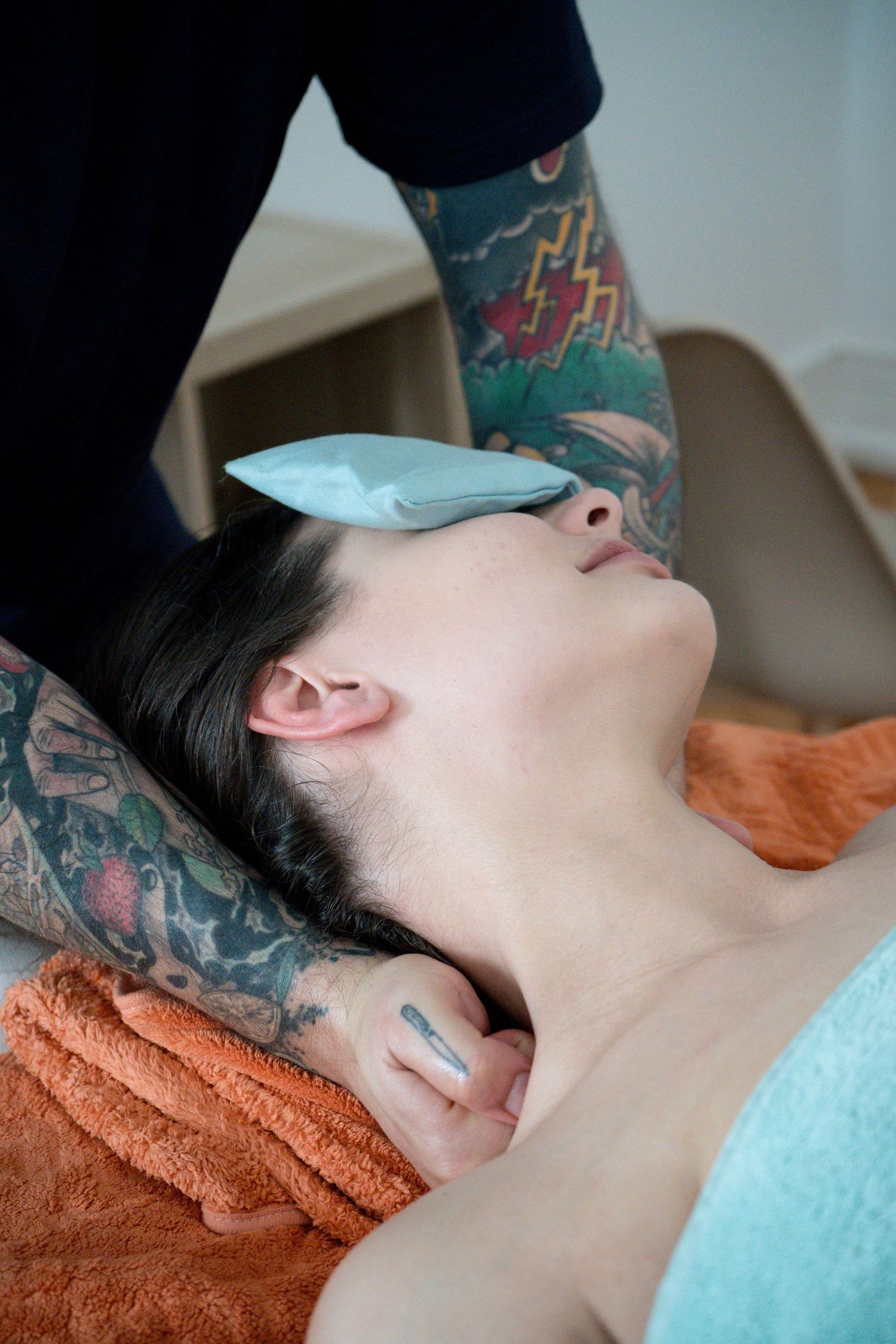 Tattooed massage therapist gently supports a woman's neck as she relaxes on orange towels with an eye pillow over her eyes.