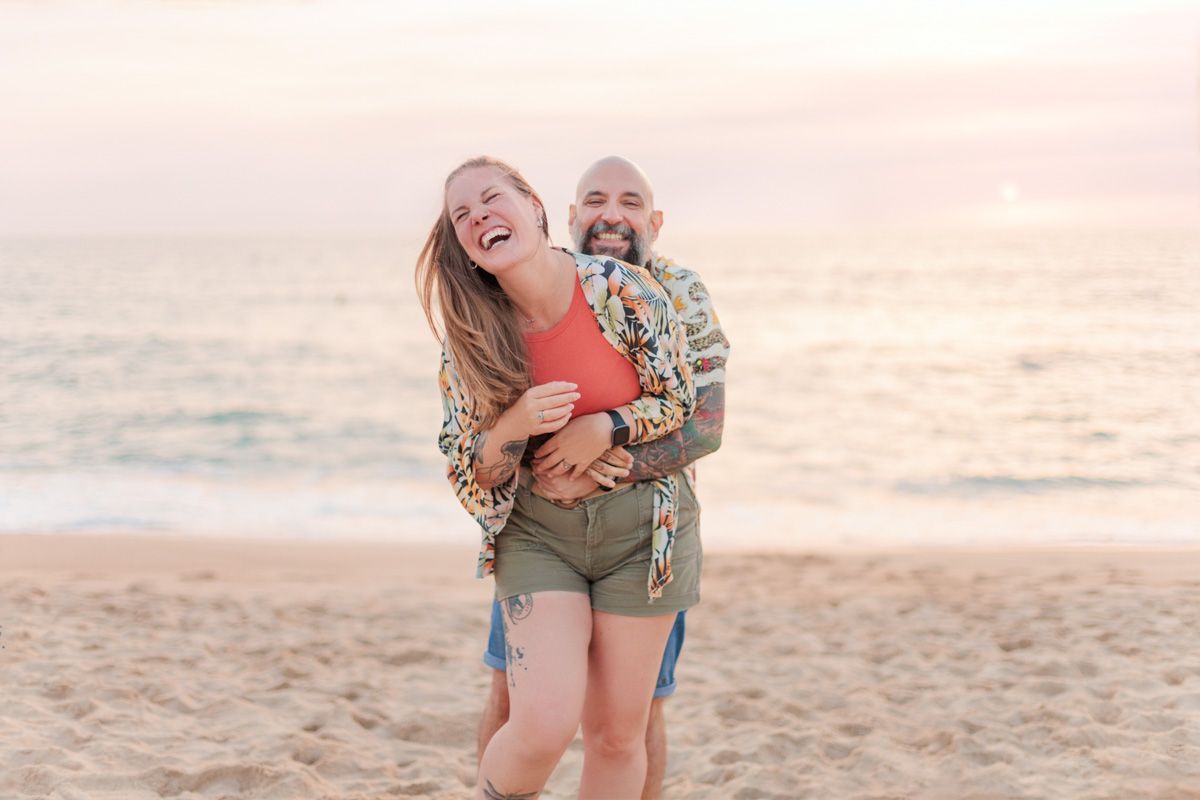 Joyful couple laughing and embracing on a sandy beach at sunset with ocean waves in the background.