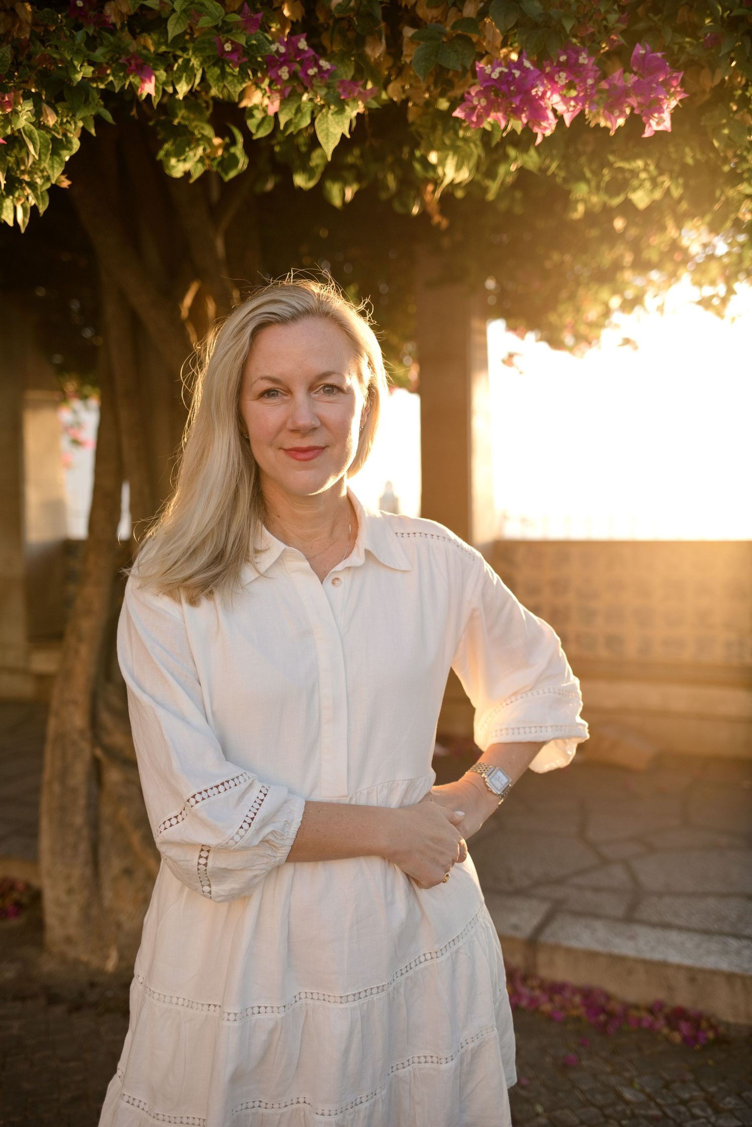 Smiling woman with long blonde hair in a white blouse and skirt stands arms crossed under a blooming bougainvillea tree at golden sunset.