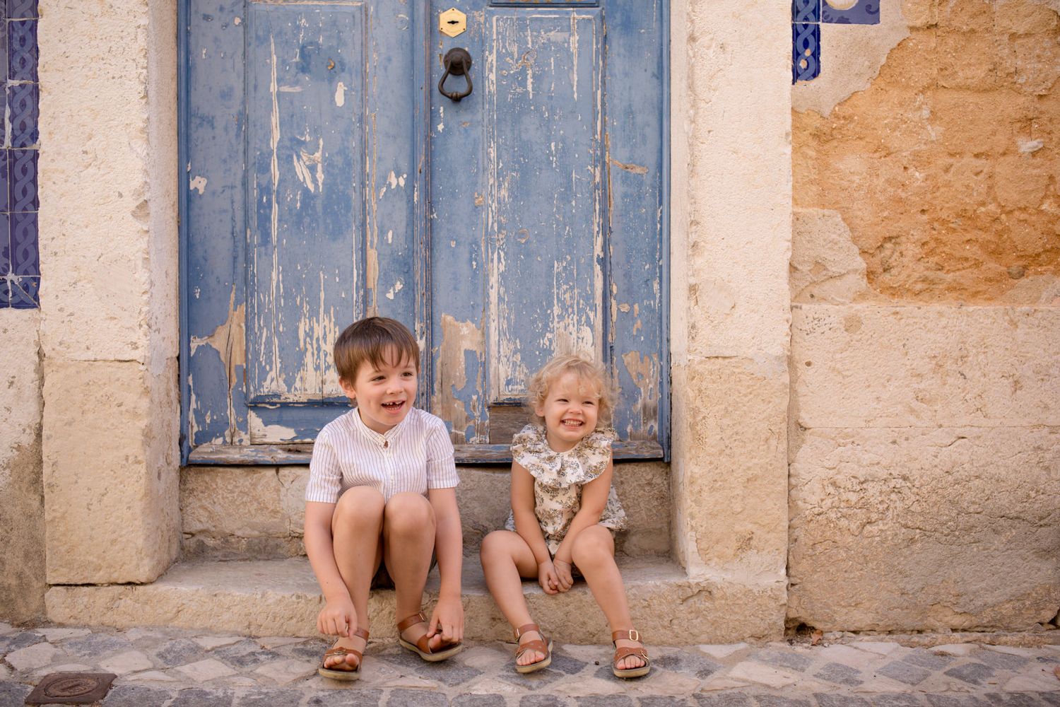 Two happy children sitting on cobblestone steps in front of a rustic blue peeling door in a charming Lisbon street.
