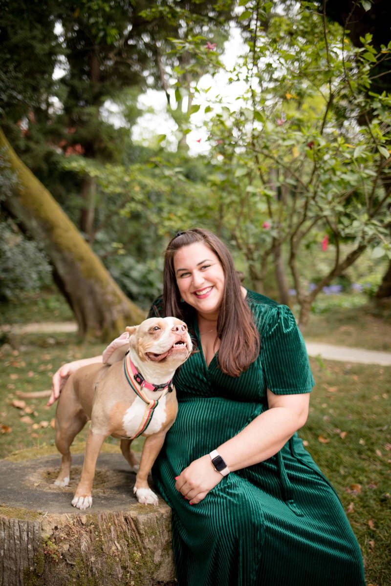 Smiling woman in green velvet dress sitting outdoors with her tan bulldog on a tree stump in a lush garden.