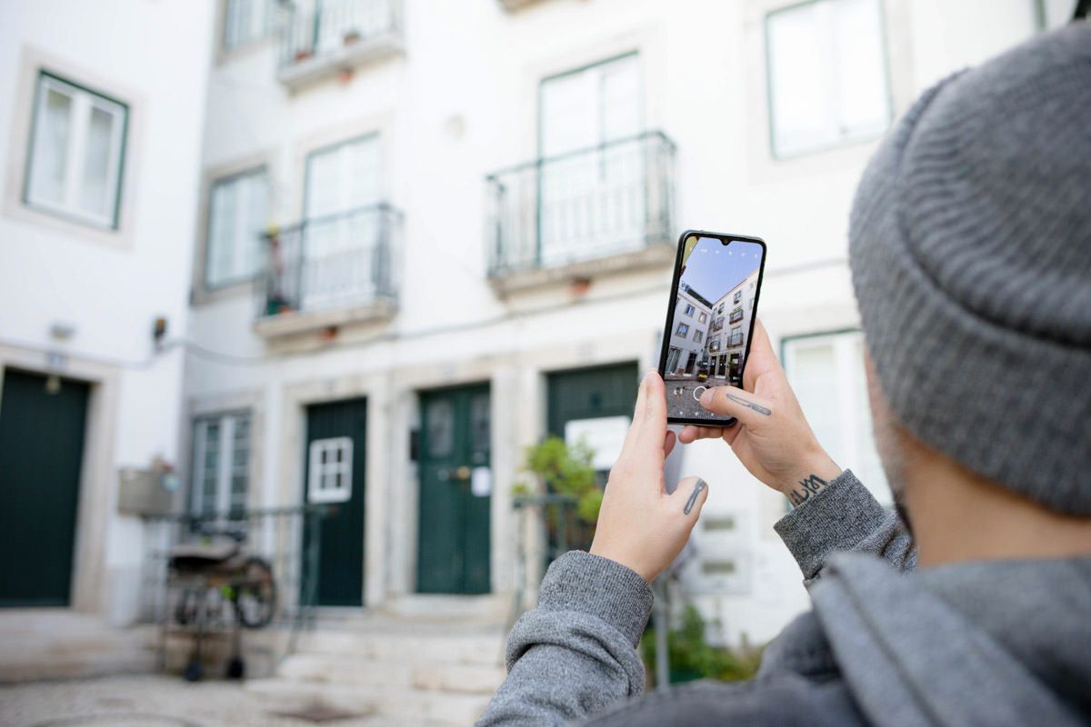 Person in gray hoodie and beanie photographing a white European building with green doors using a smartphone.