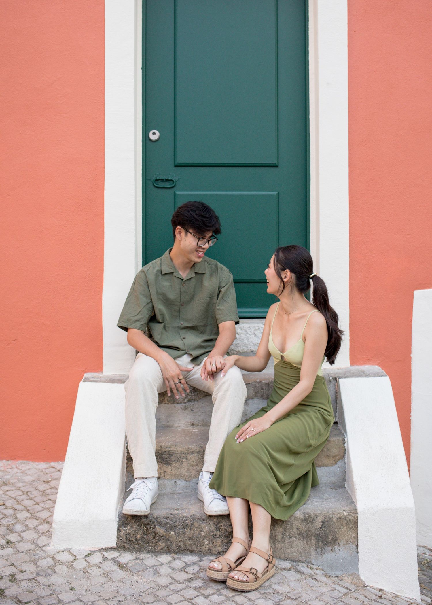 A couple sits on steps outside a coral building with a green door, smiling at each other in matching green outfits.