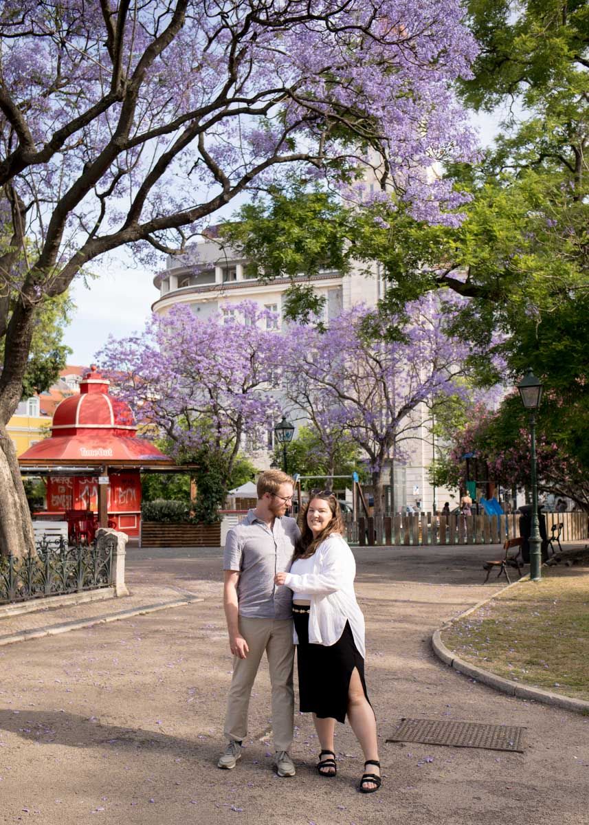 Couple embracing under blooming purple jacaranda trees in a scenic park with historic buildings.