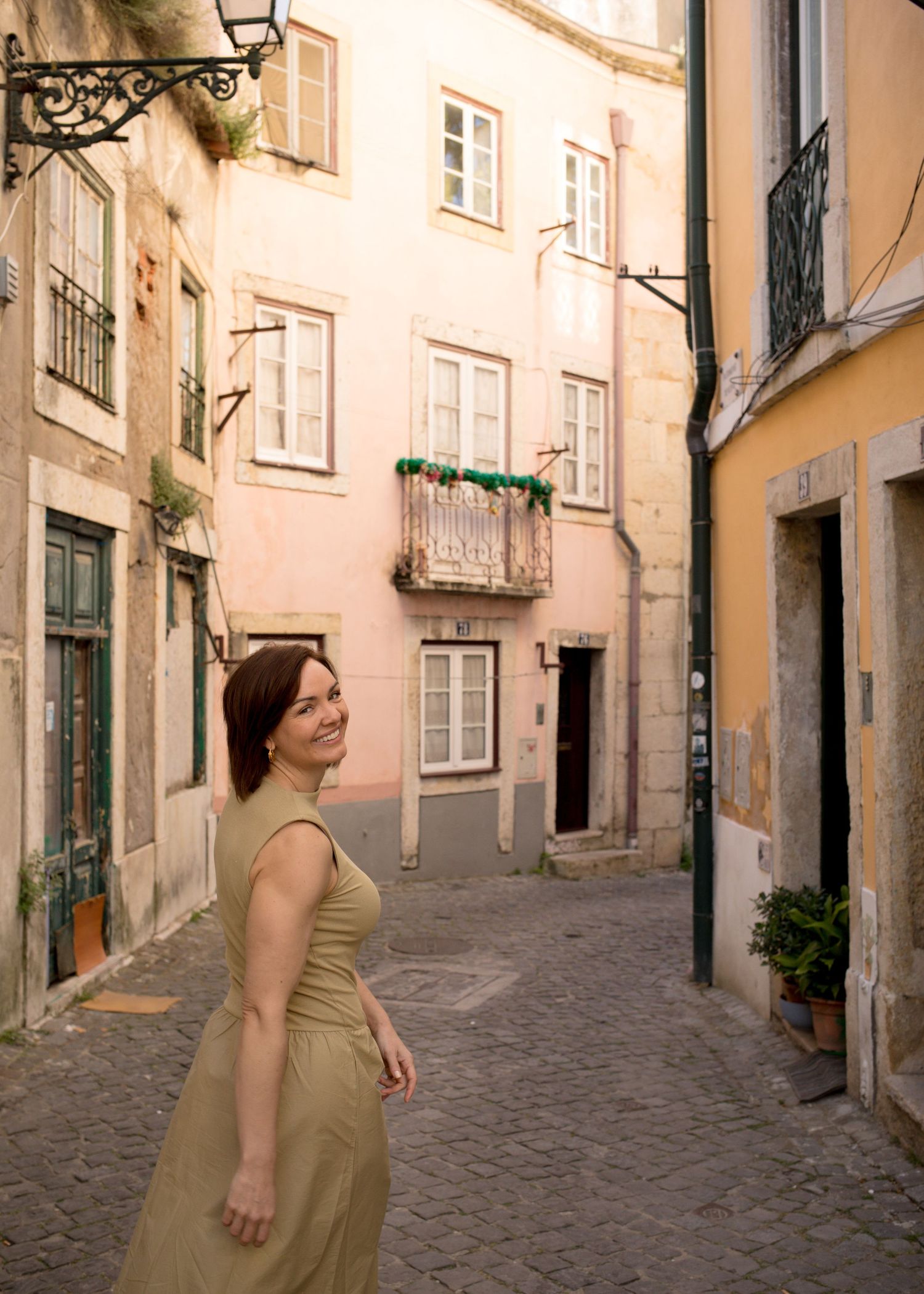 Smiling woman in olive dress turning in charming cobblestone alley with pastel buildings.