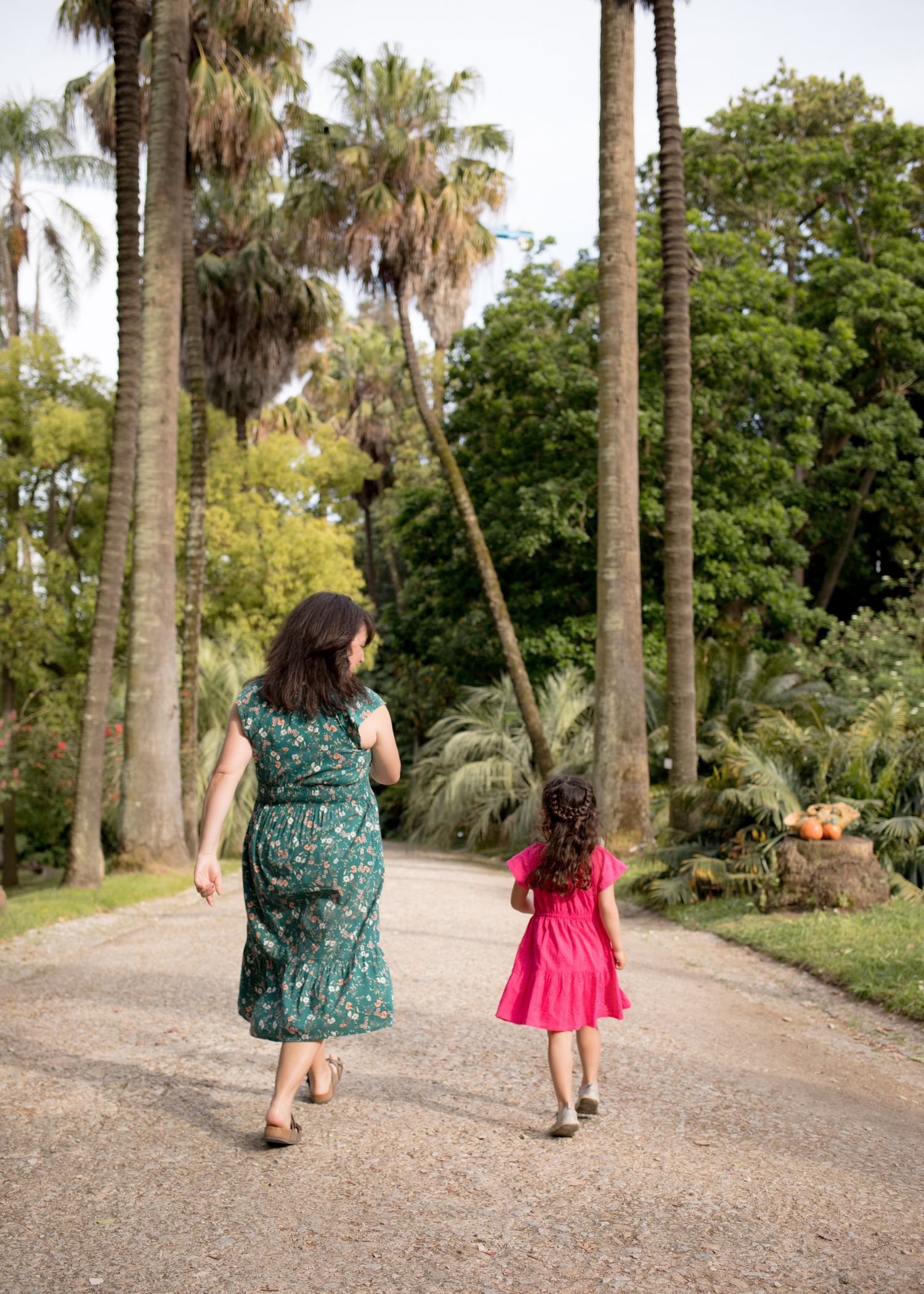 Woman in green floral dress walking with young girl in pink dress along a palm tree-lined path.