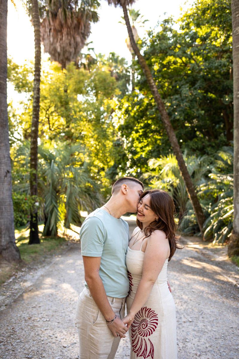 Couple sharing a tender kiss on a sunlit garden path surrounded by lush green palm trees.