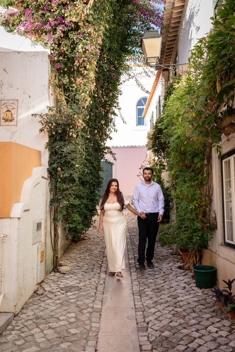 Couple walking hand-in-hand down a charming cobblestone alley lined with ivy and blooming flowers in a European village.