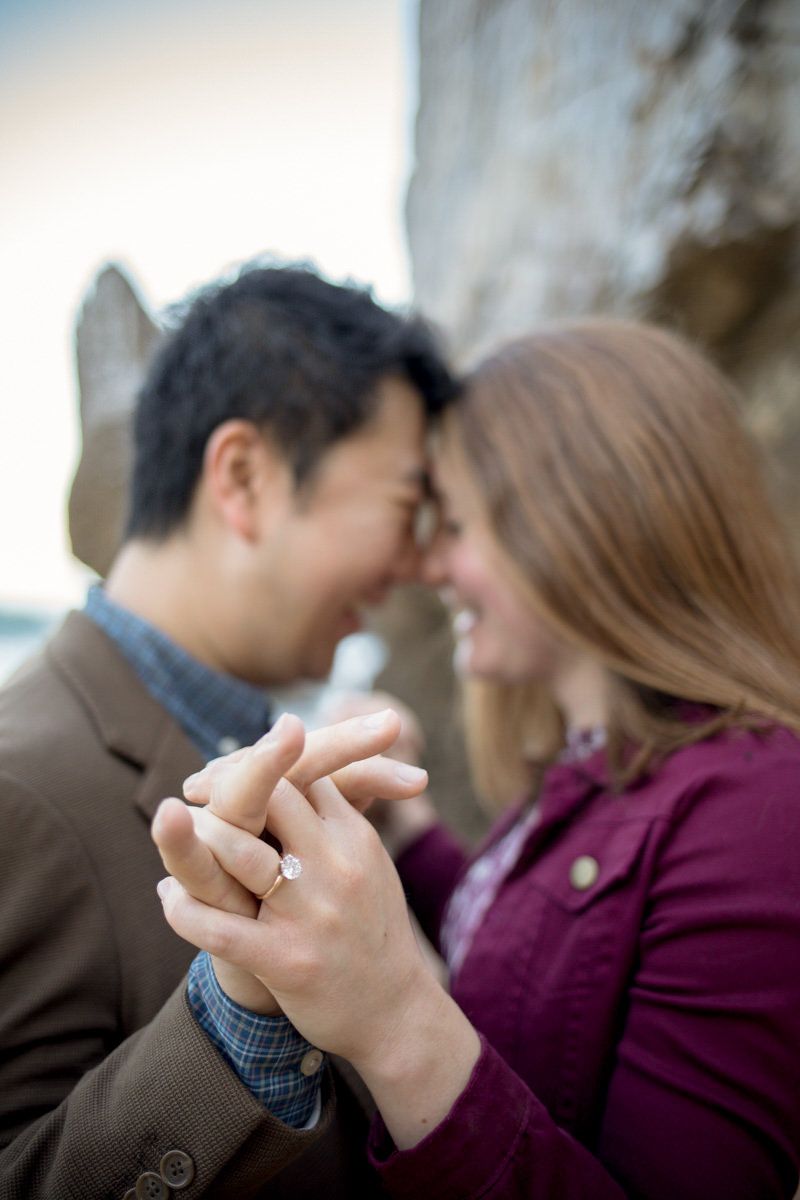 A joyful engaged couple holds hands and touches foreheads, showcasing a diamond engagement ring outdoors.