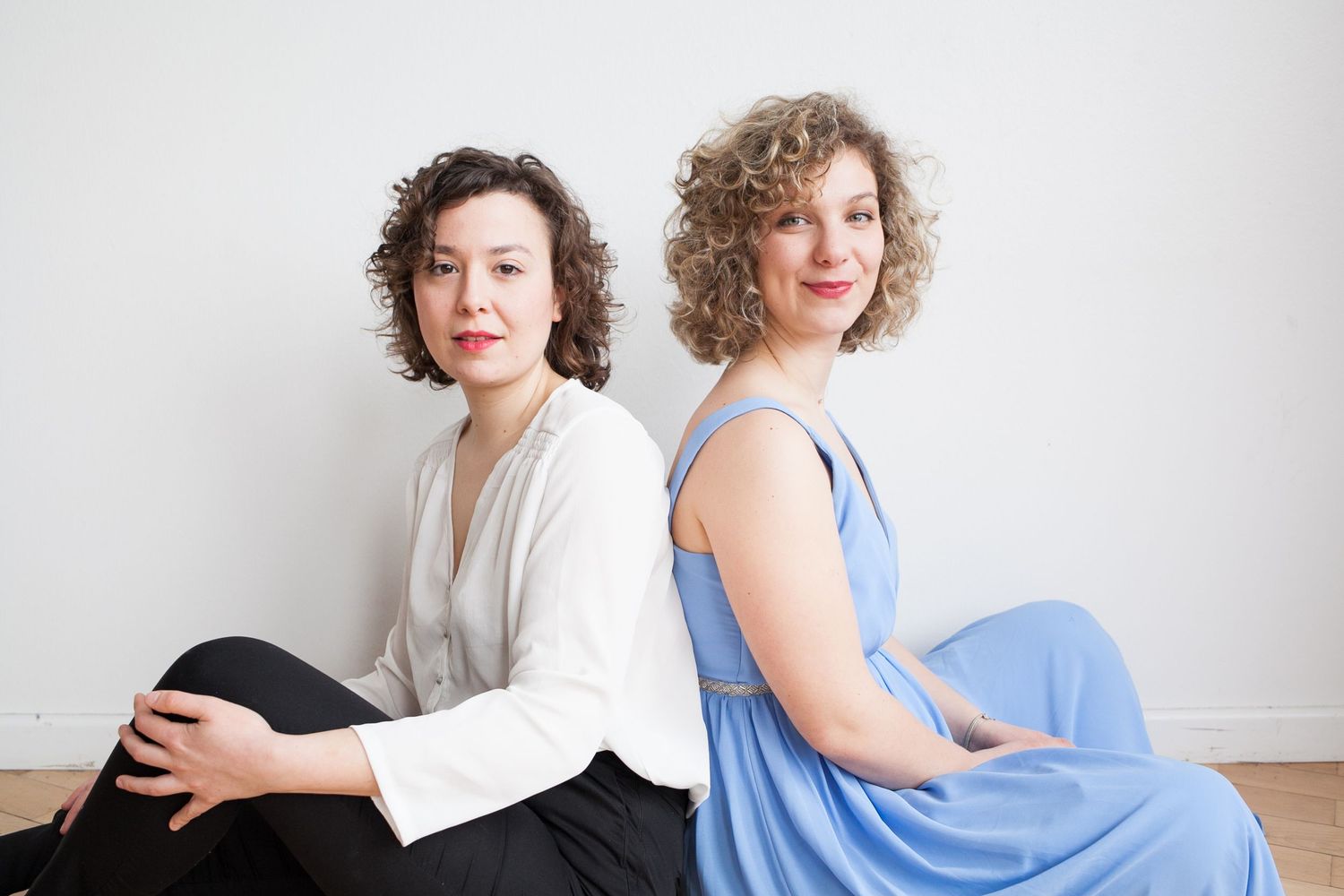 Two women with curly hair sit back to back on a wooden floor, one in a white blouse and black pants, the other in a light blue dress.
