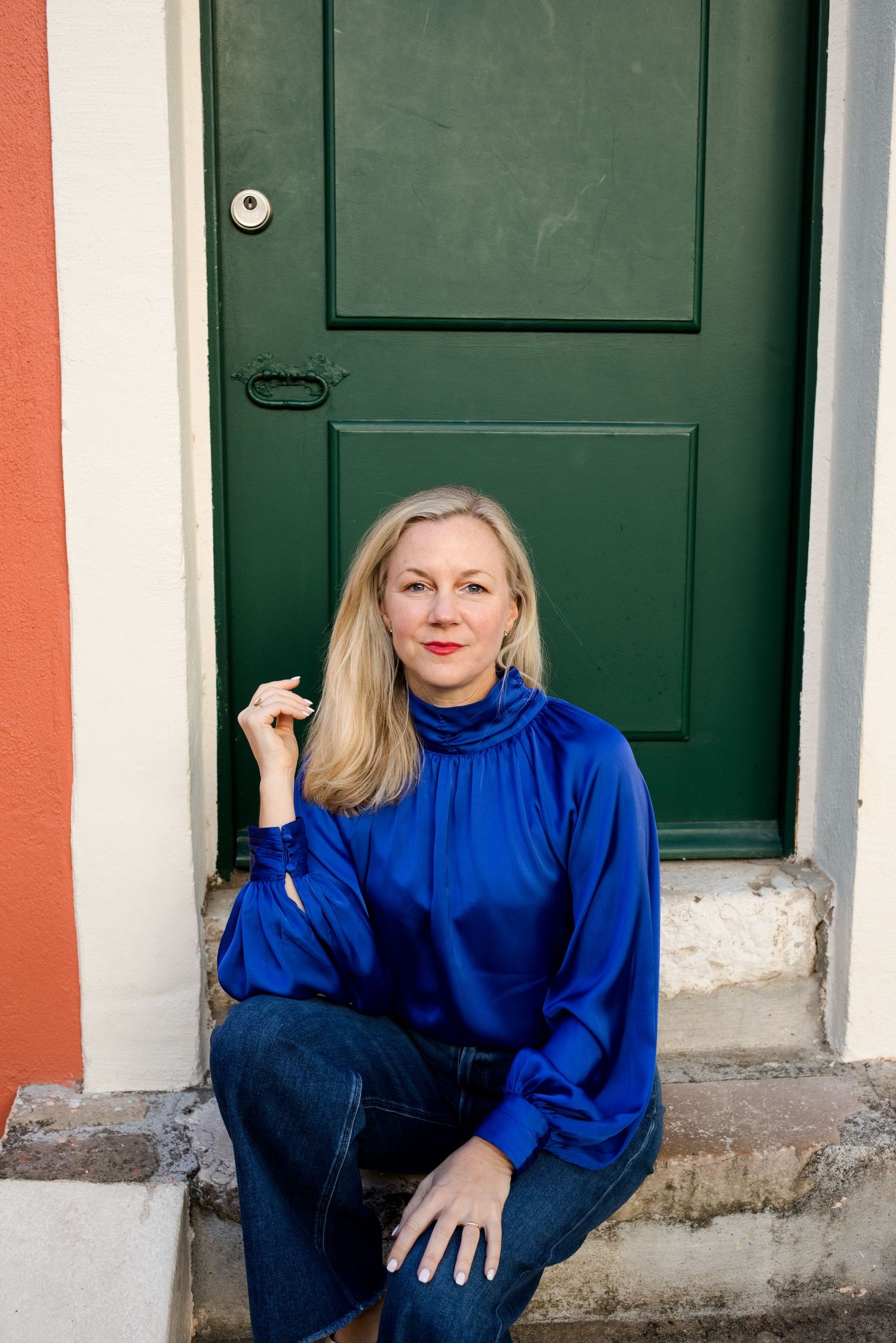 Woman with long blonde hair in a royal blue satin blouse and jeans sits on a stone step in front of a green wooden door.