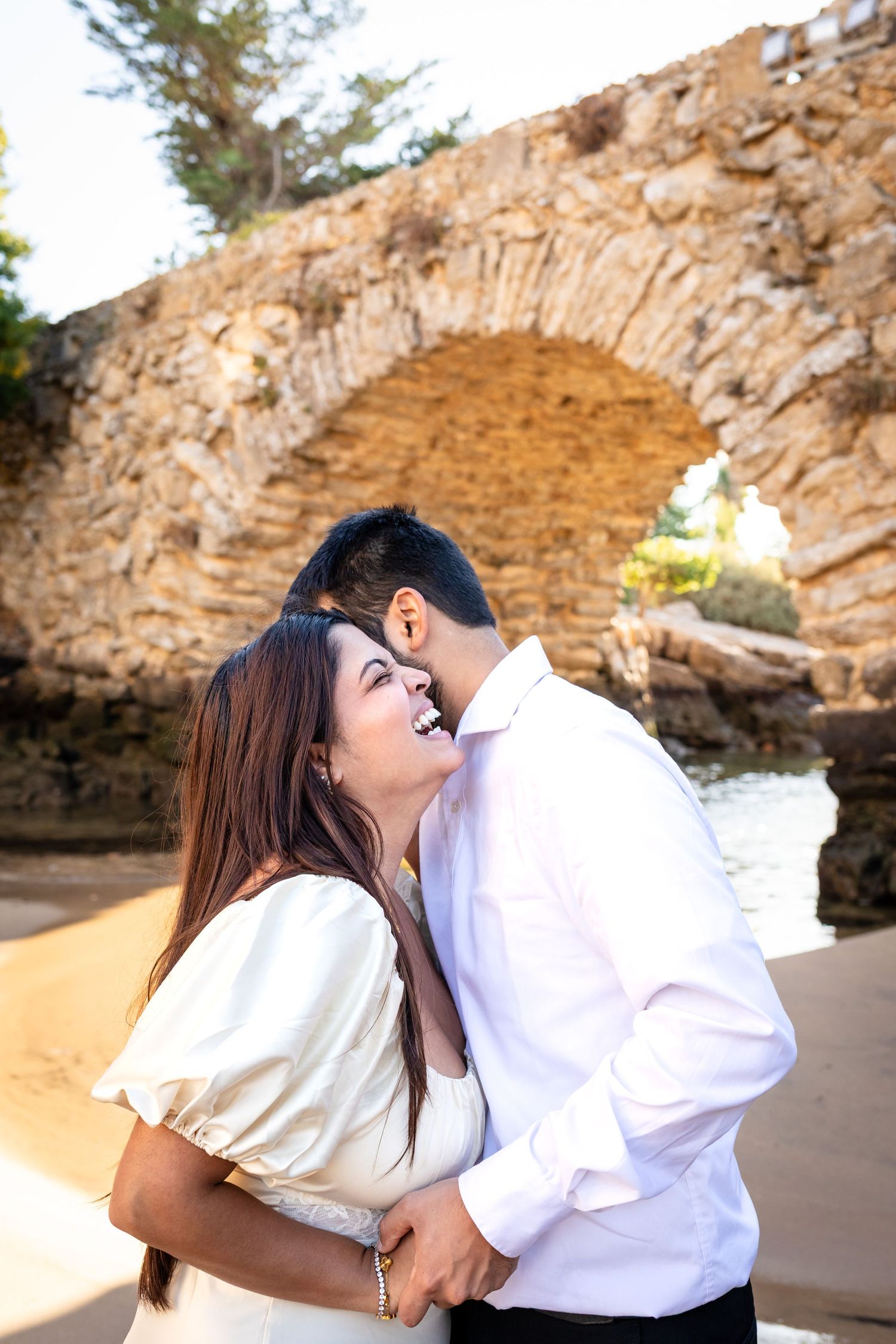 Couple laughing beneath a stone arch after a romantic Cascais proposal photoshoot by the beach.