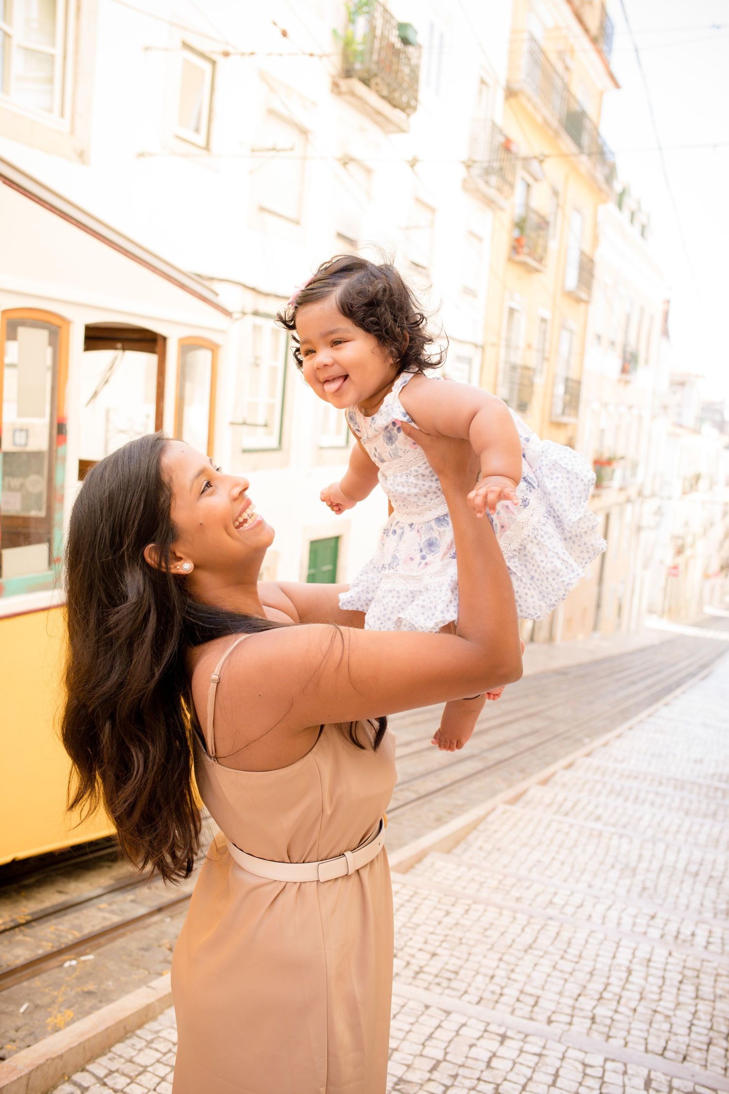 Joyful mother lifts laughing baby girl in white dress on a cobblestone street with yellow tram in Lisbon.