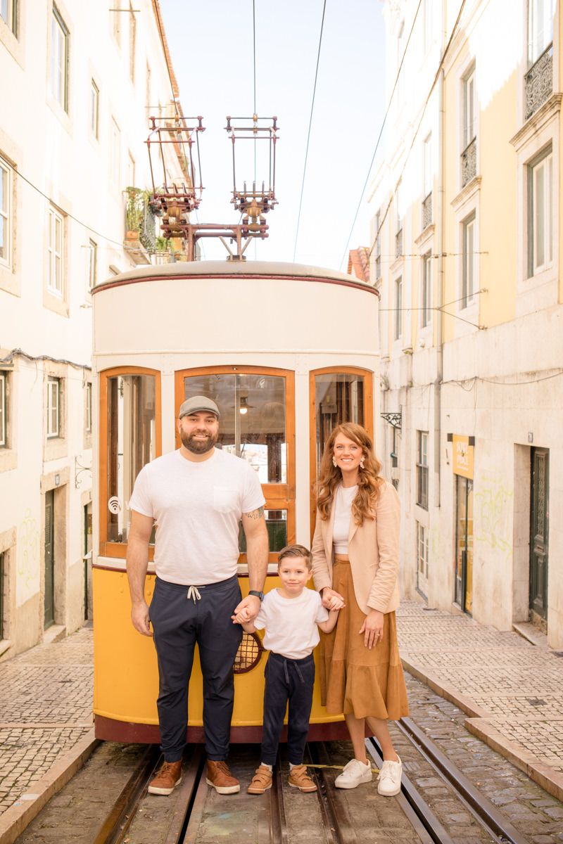 Family of three posing in front of a yellow vintage tram on a narrow cobblestone street in Lisbon, Portugal.