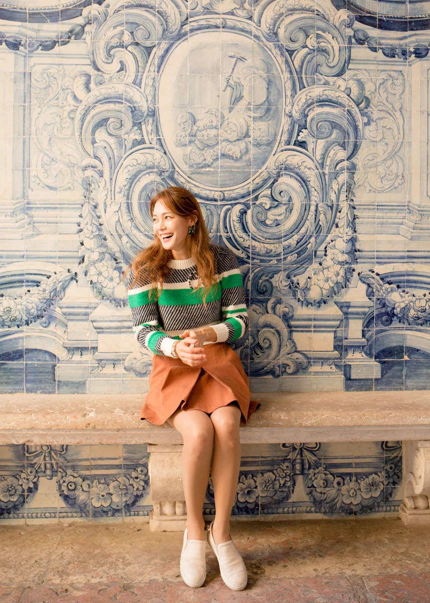 Woman in striped sweater sits laughing before ornate blue and white Portuguese azulejo tile wall.
