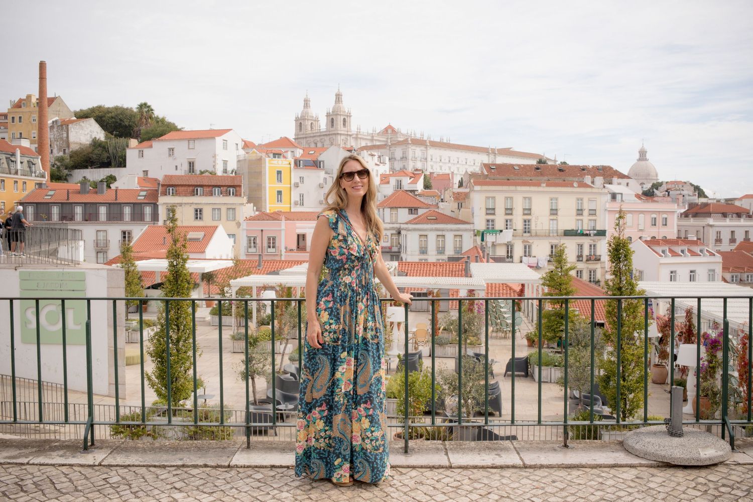 Woman in floral maxi dress standing at viewpoint overlooking Lisbon cityscape.