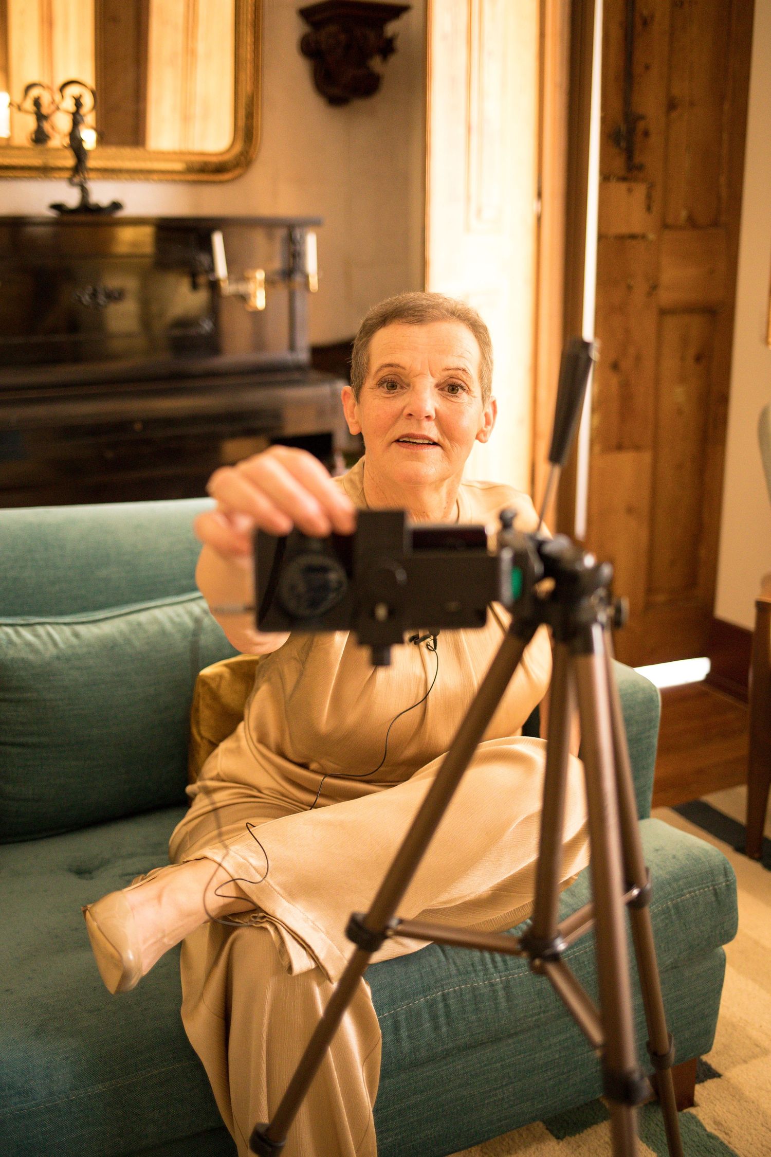 Woman in a satin beige outfit sits on a green sofa adjusting a camera mounted on a wooden tripod in a warmly lit room.