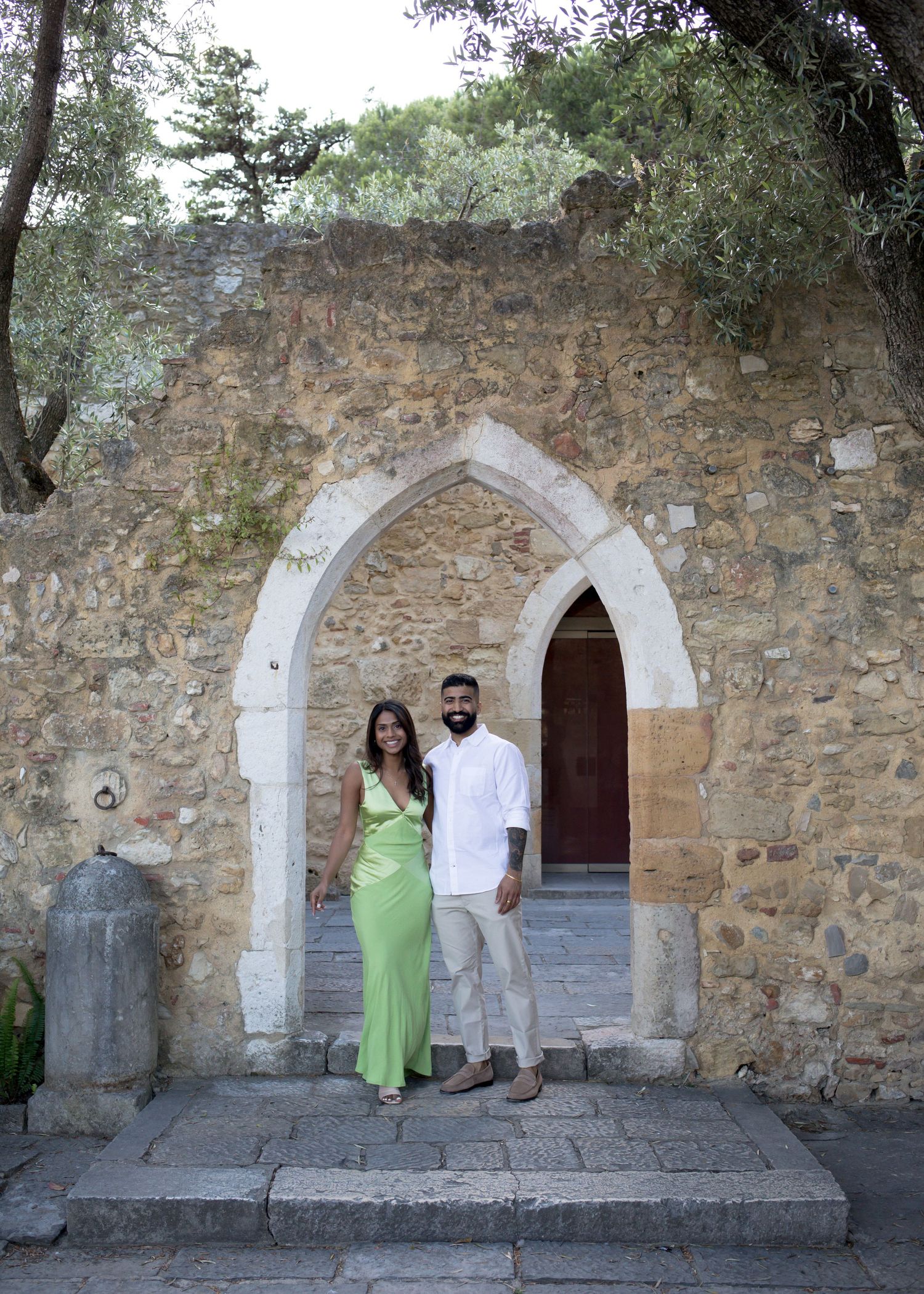Couple posing under a Gothic stone arch at a historic Mediterranean landmark, woman in green dress, man in white shirt.