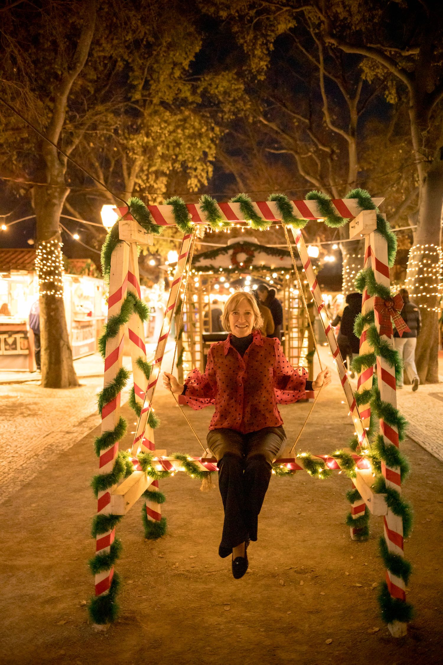 Woman smiling on a festive Christmas swing decorated with candy cane stripes and green garland lights at a holiday event.
