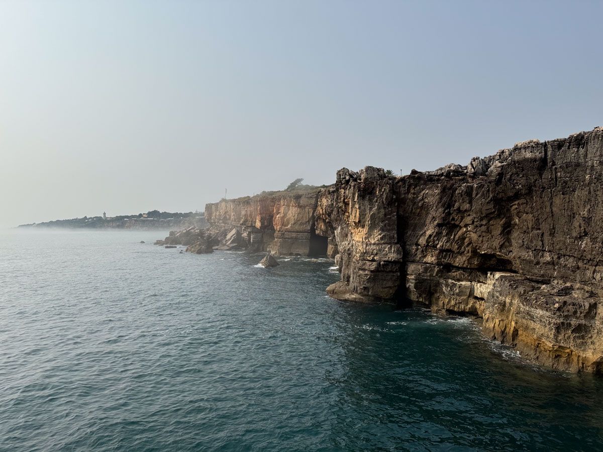 Rocky coastal cliffs overlooking calm blue ocean waters under a hazy sky, showcasing dramatic natural rock formations.