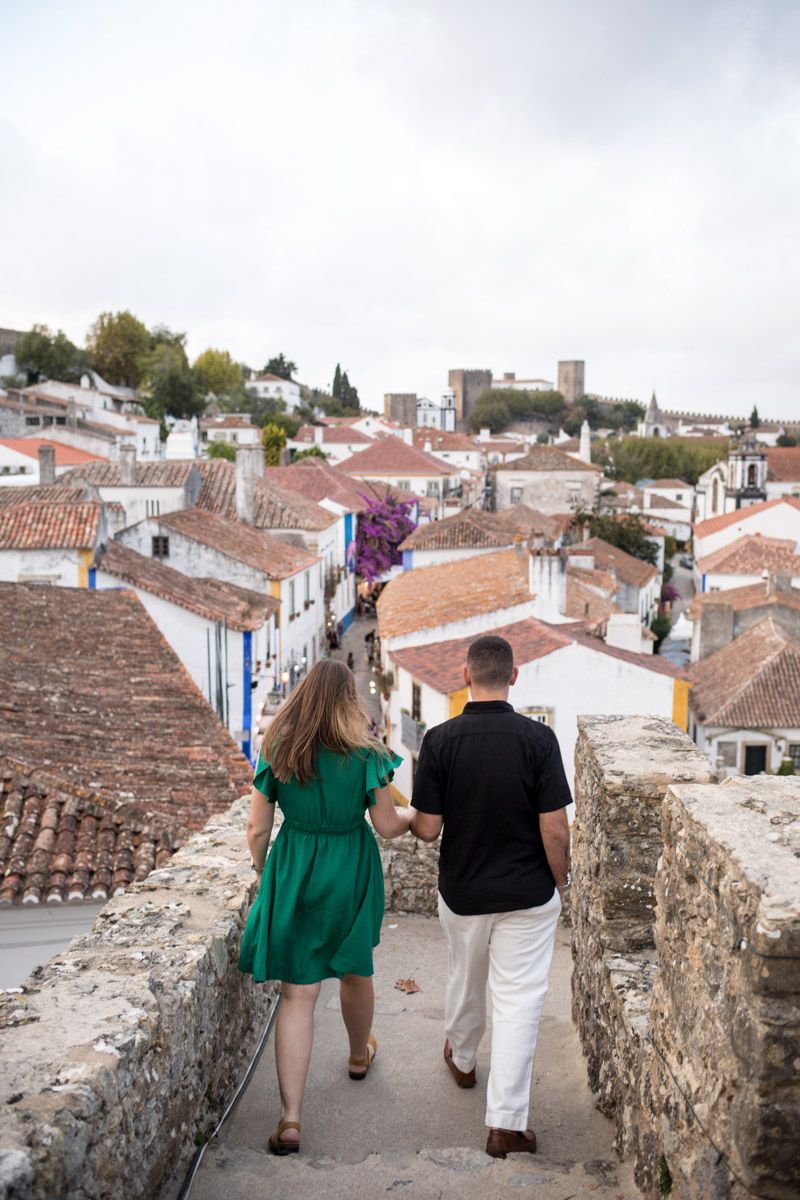 Couple holding hands walking along stone wall overlooking white-washed village rooftops in Óbidos, Portugal.