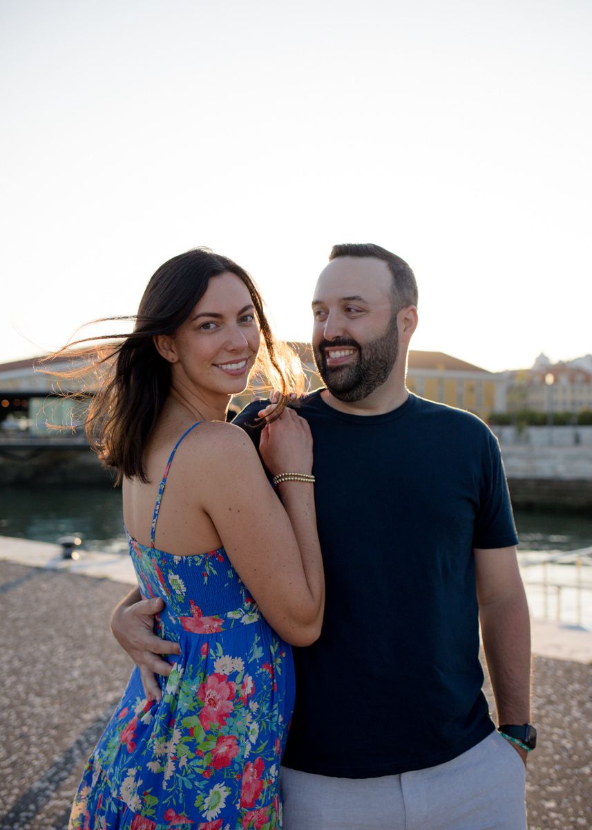 A smiling couple cuddle beside the water in Lisbon during golden hour, with soft sunlight behind them and city buildings in the background.