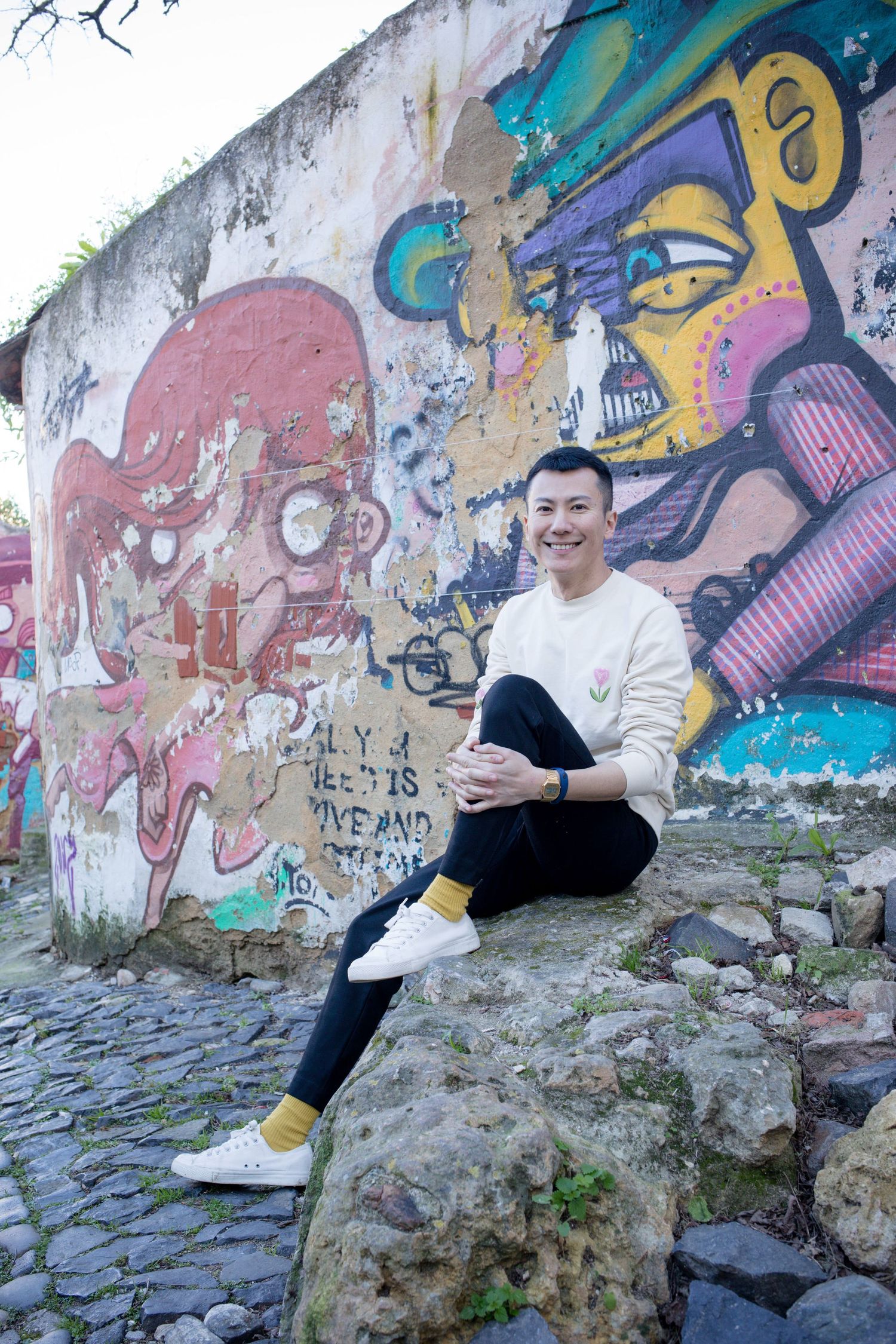 Smiling person in cream sweater sits on stone wall before colorful street art graffiti mural.
