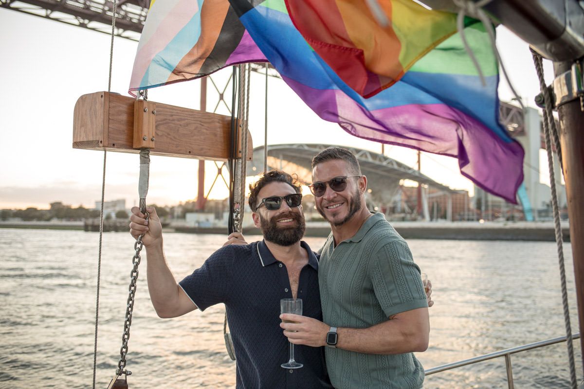 Two men smiling on a sailboat with a Pride rainbow flag flying behind them at sunset near a harbor bridge.