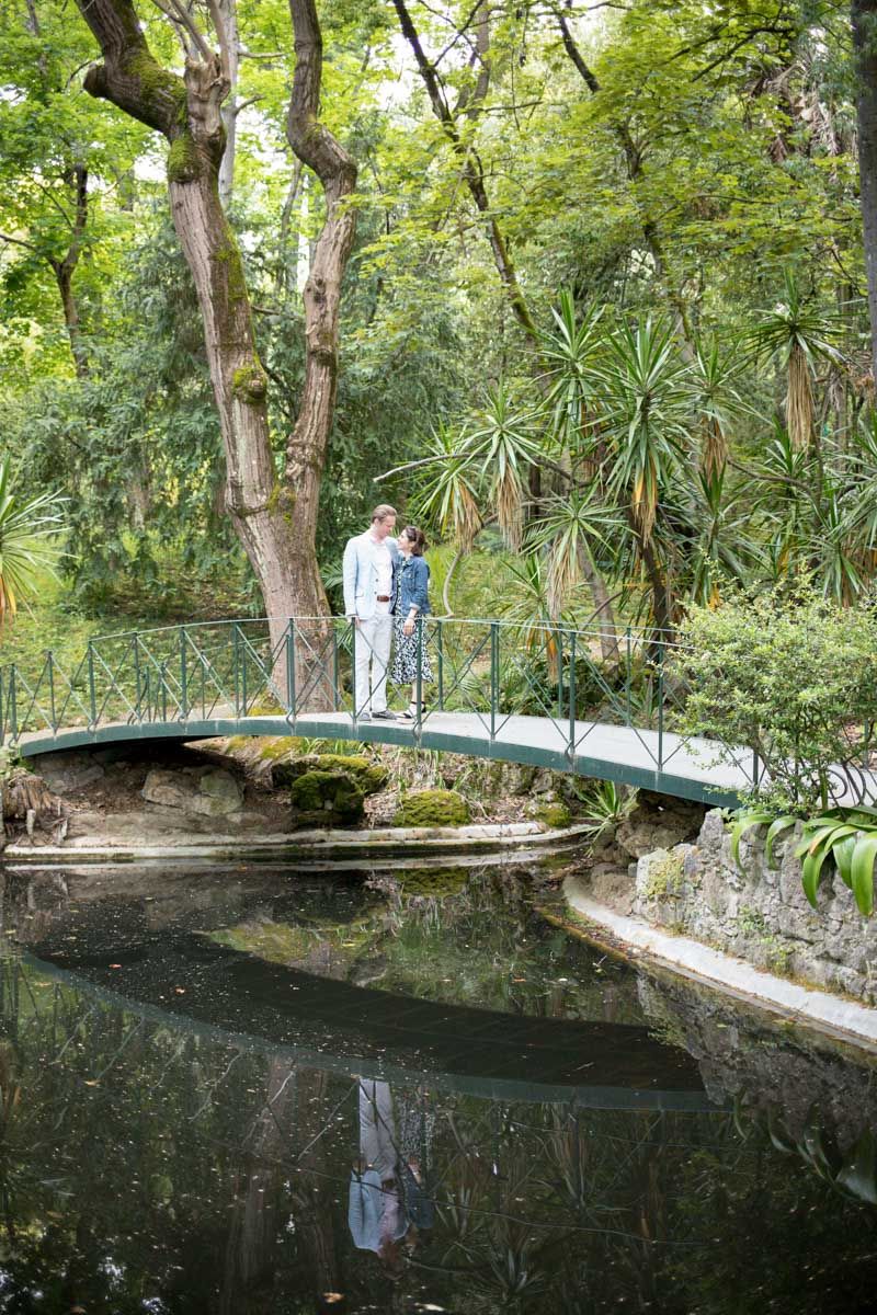 Couple standing on ornate green bridge over calm pond surrounded by lush tropical garden trees and plants.