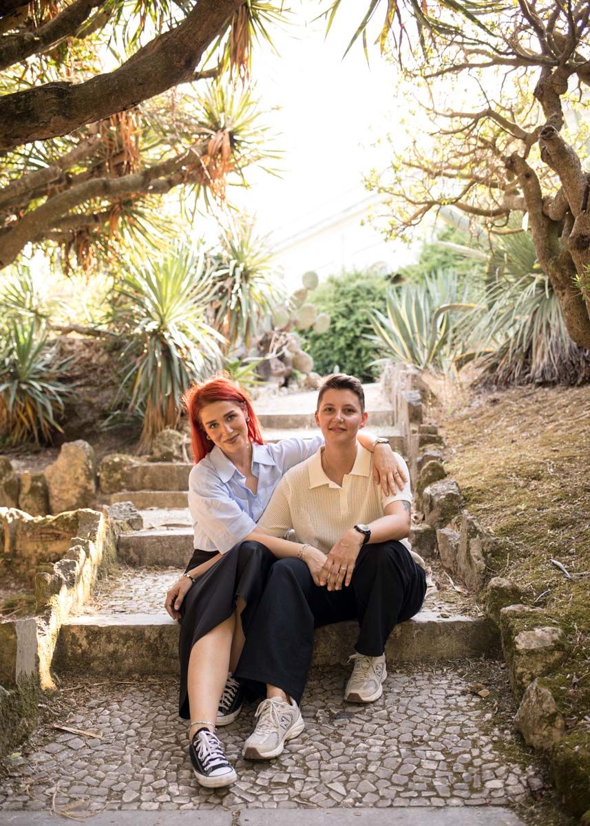 Two women sitting together on stone steps in a lush garden, one with red hair wearing a blue shirt.