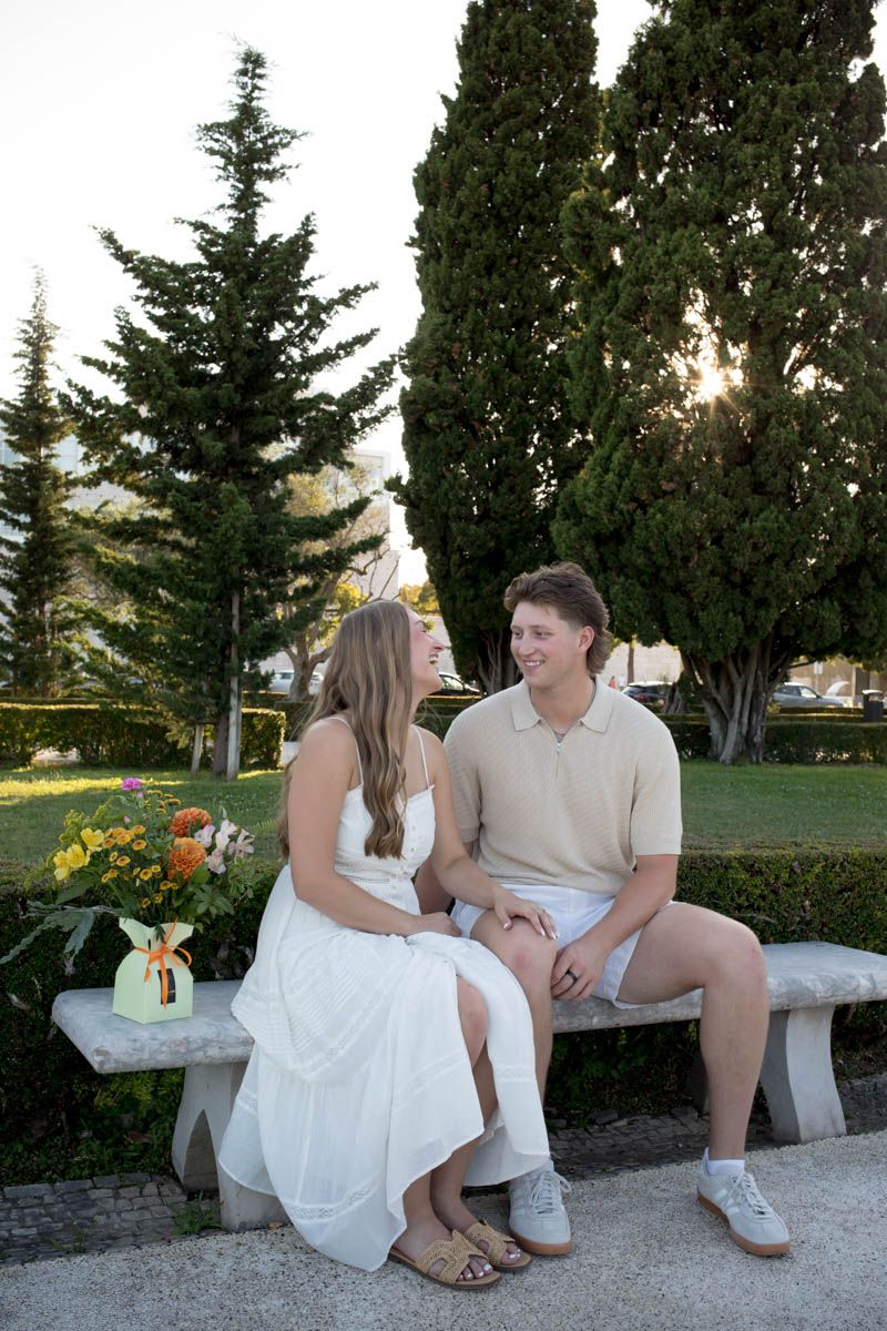 A couple sits on a stone bench in a park, holding hands and smiling at each other, surrounded by tall green trees.