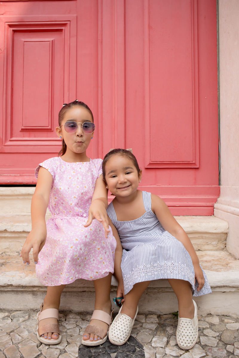 Two young girls in summer dresses sitting on steps in front of a red door, one wearing sunglasses, during a photoshoot in Lisbon.