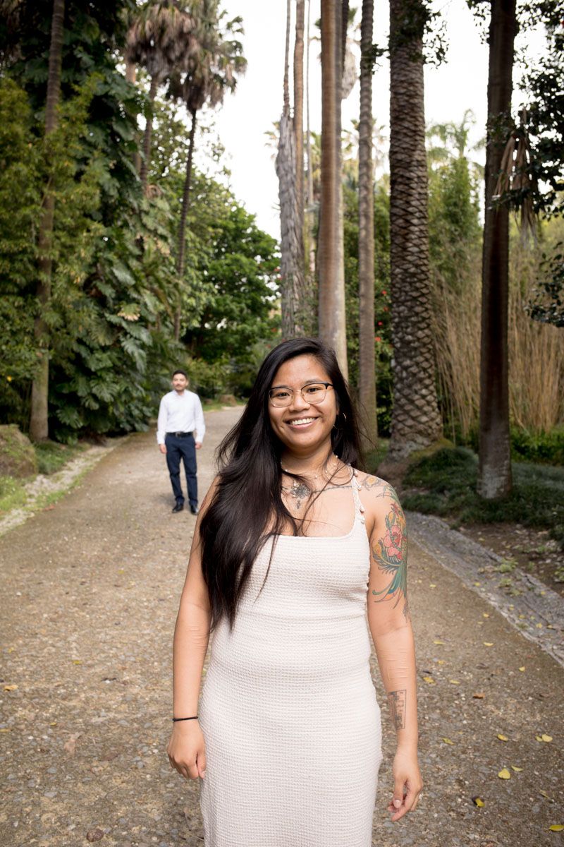 Smiling tattooed woman in white dress on palm-lined path with man walking behind her.