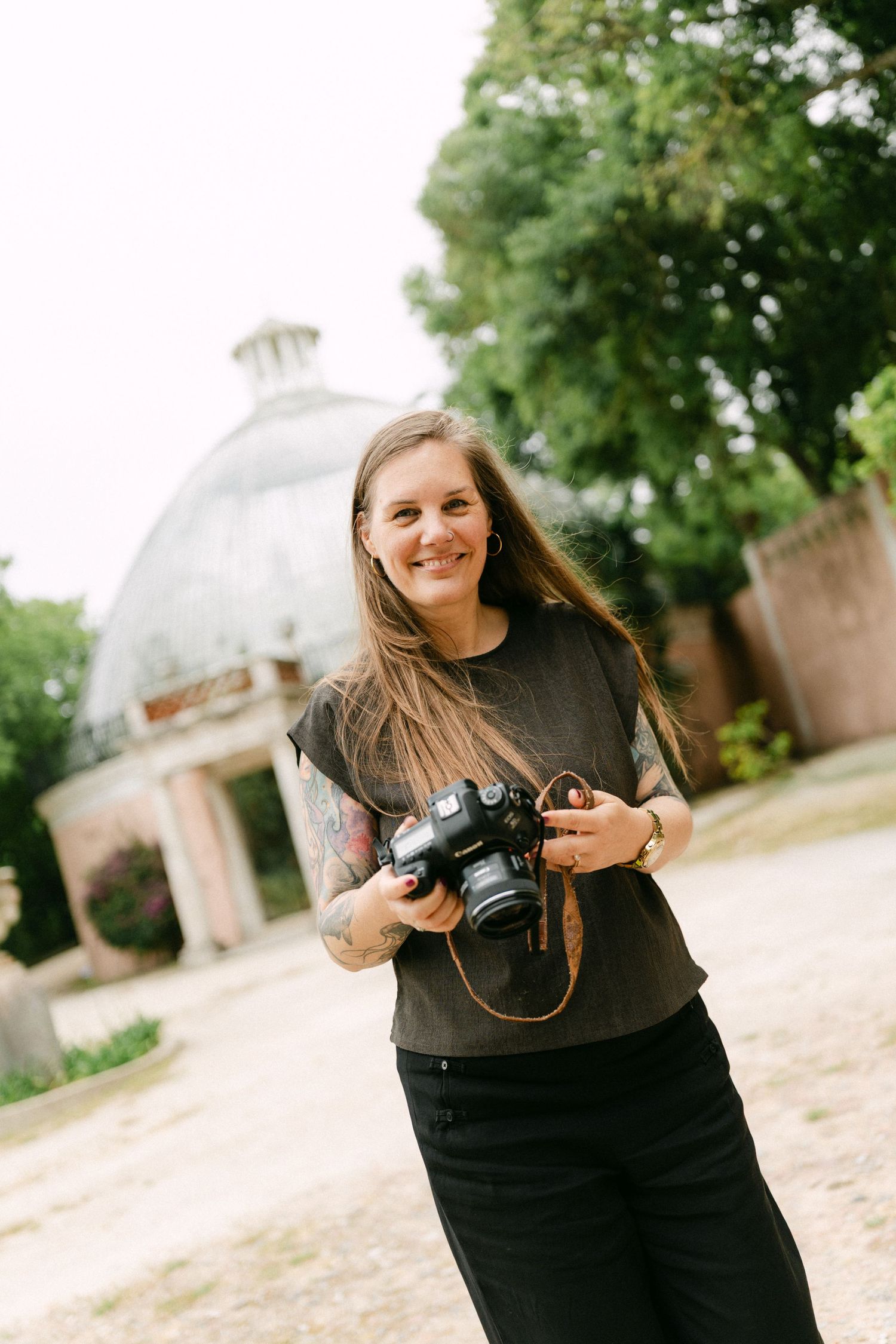 Smiling woman with tattoos holding a DSLR camera outdoors near a historic domed building surrounded by trees.