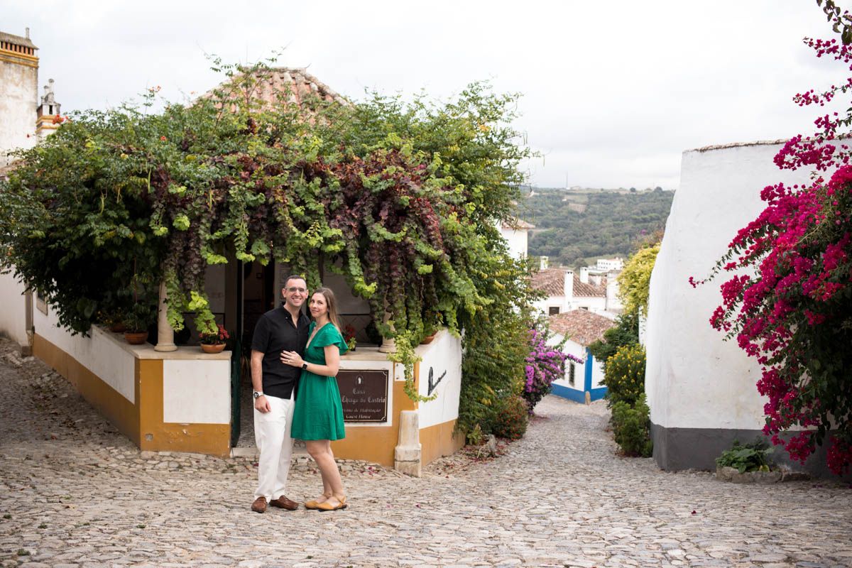 Couple embracing on cobblestone street in Óbidos, Portugal, surrounded by lush greenery and colorful bougainvillea.
